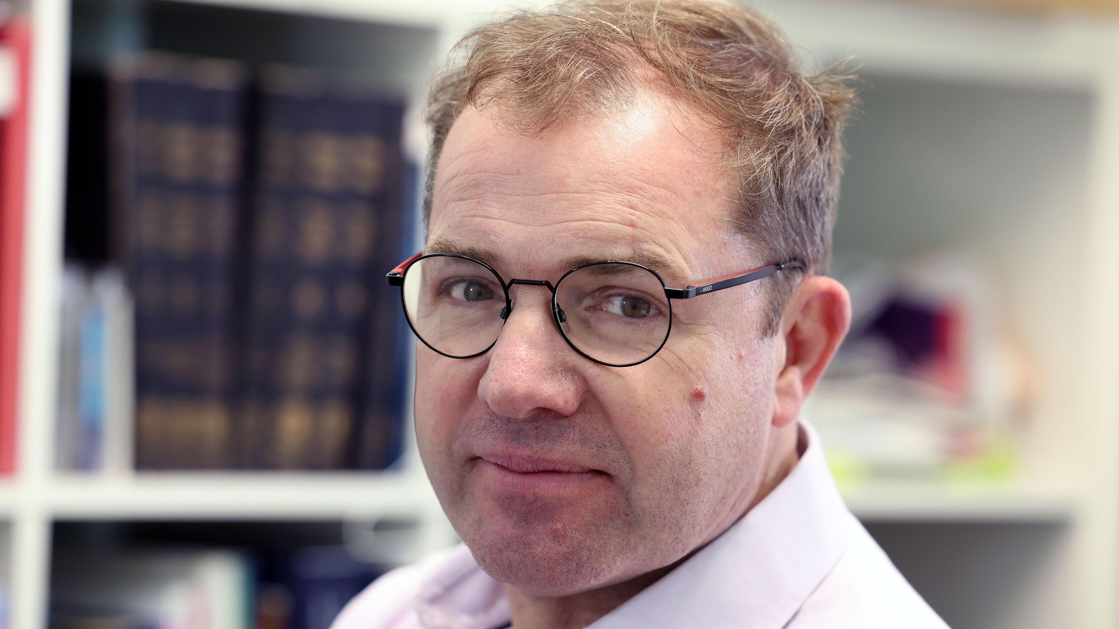 Prof Torkington is wearing glasses and a pale purple shirt. In the background are books on a shelf.