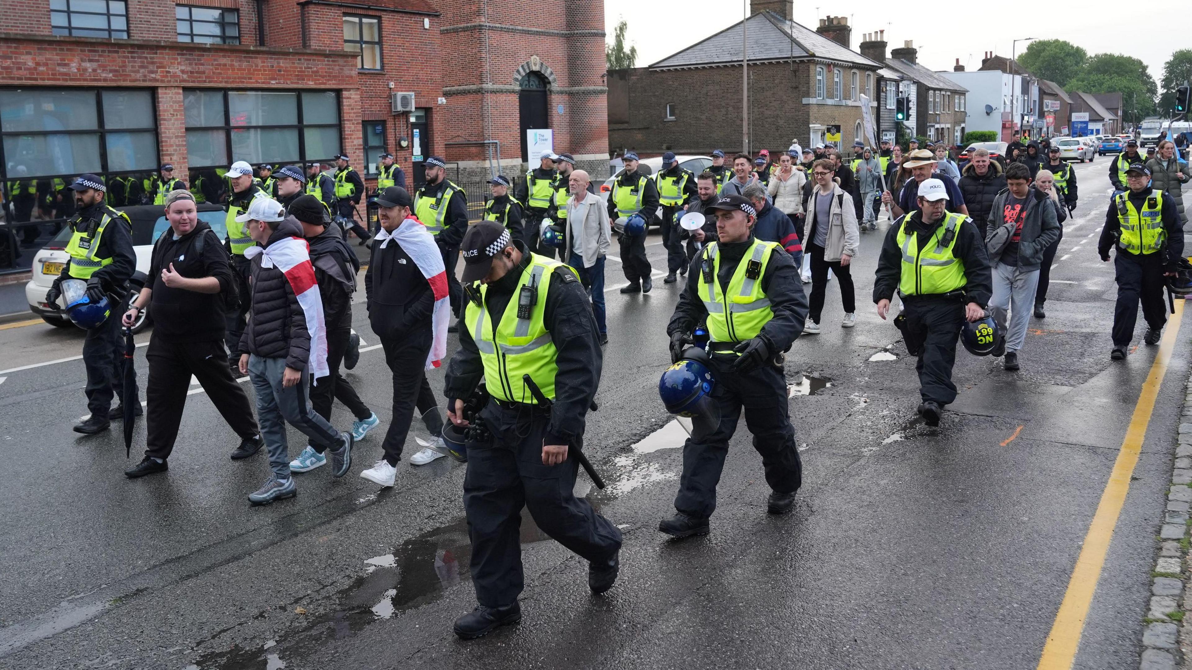 Police are walking alongside protesters on a road near the Bell Hotel in Epping. Some people can be seen with England flags over their shoulders