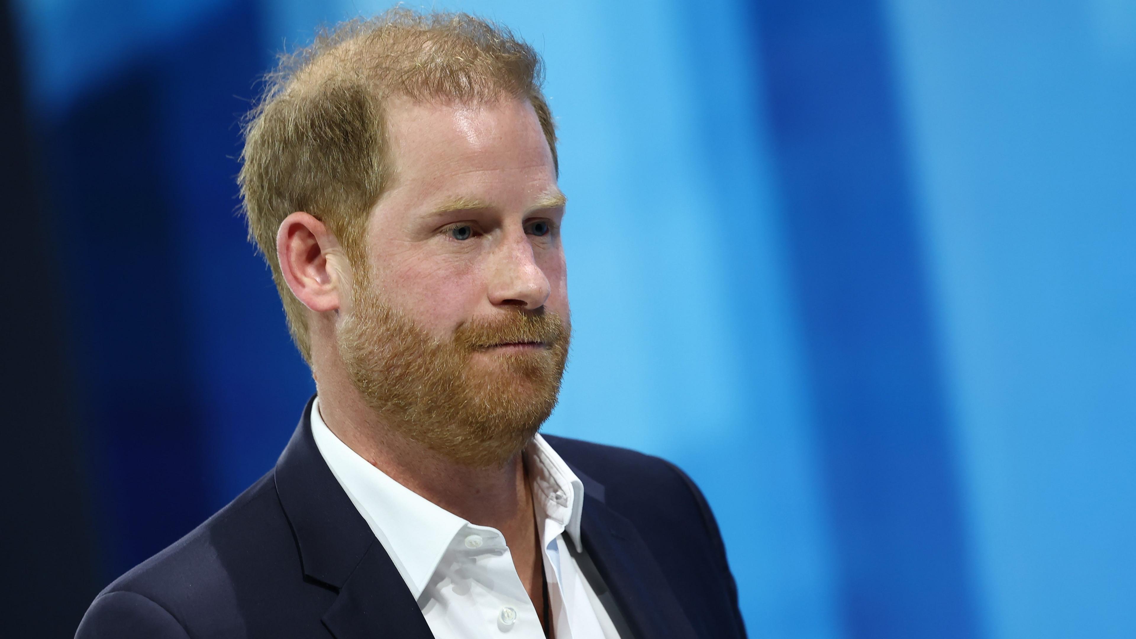 File photo of Prince Harry on a blue background, wearing a white open-neck shirt and a dark blazer.