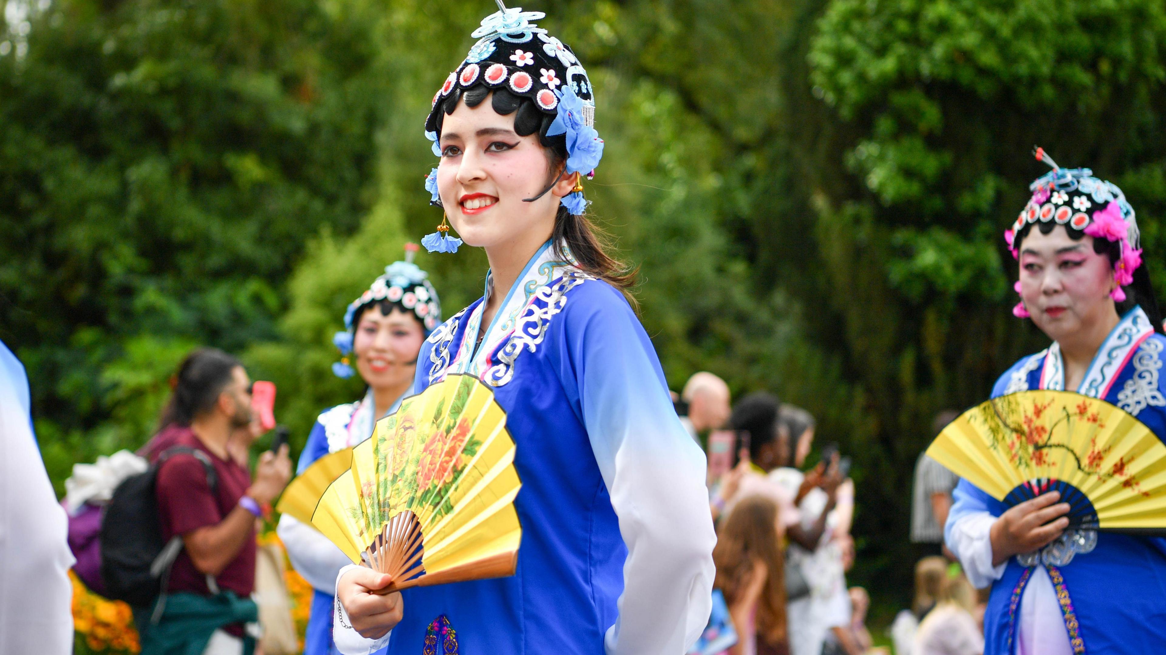 Women are holding fans that are decorated with flowers. They're wearing blue and taking part in a festival. Crowds are watching.