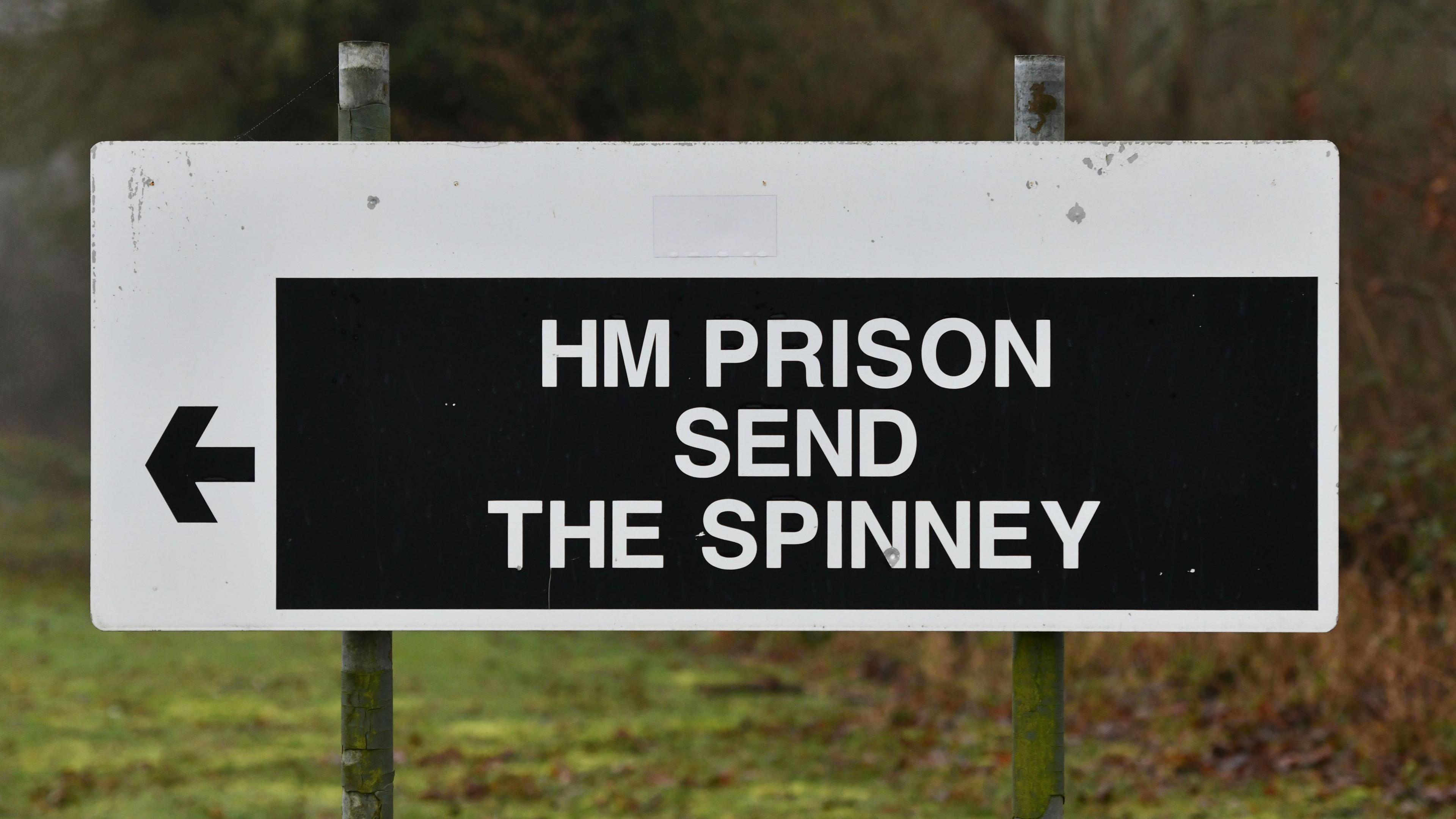 A sign reading HM PRISON SEND THEN SPINNEY written in white lettering with a black arrow pointing to the left