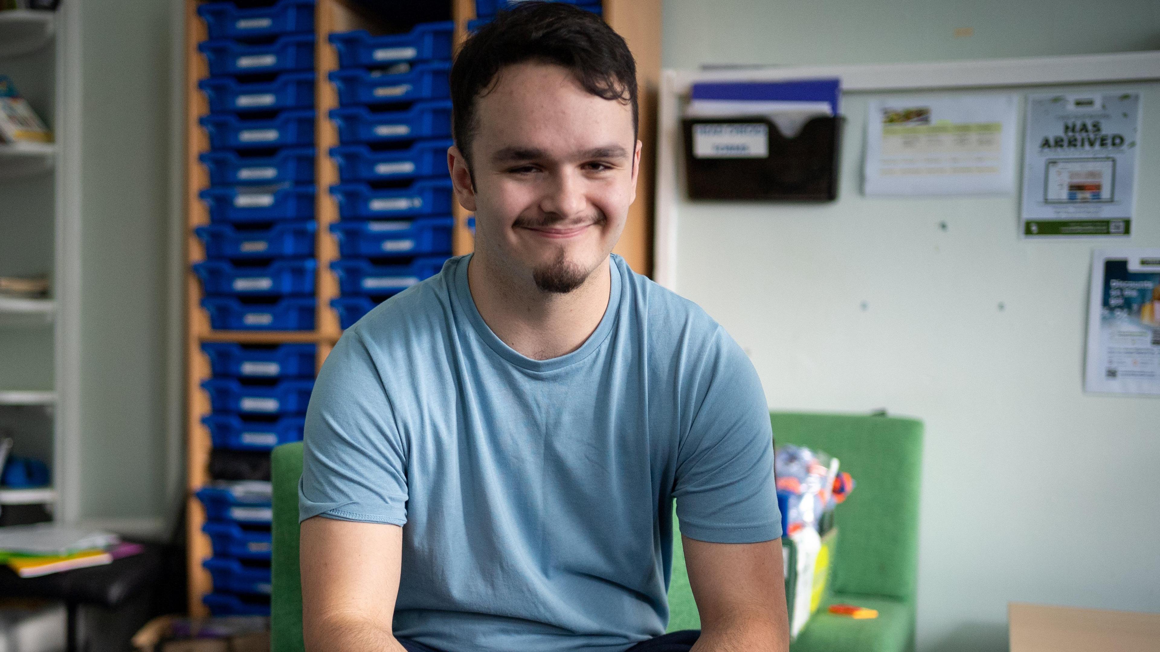 Harry smiling and looking straight at the camera, wearing a blue T-shirt, in a classroom with shelves behind him. He has short dark hair and a goatee beard. 