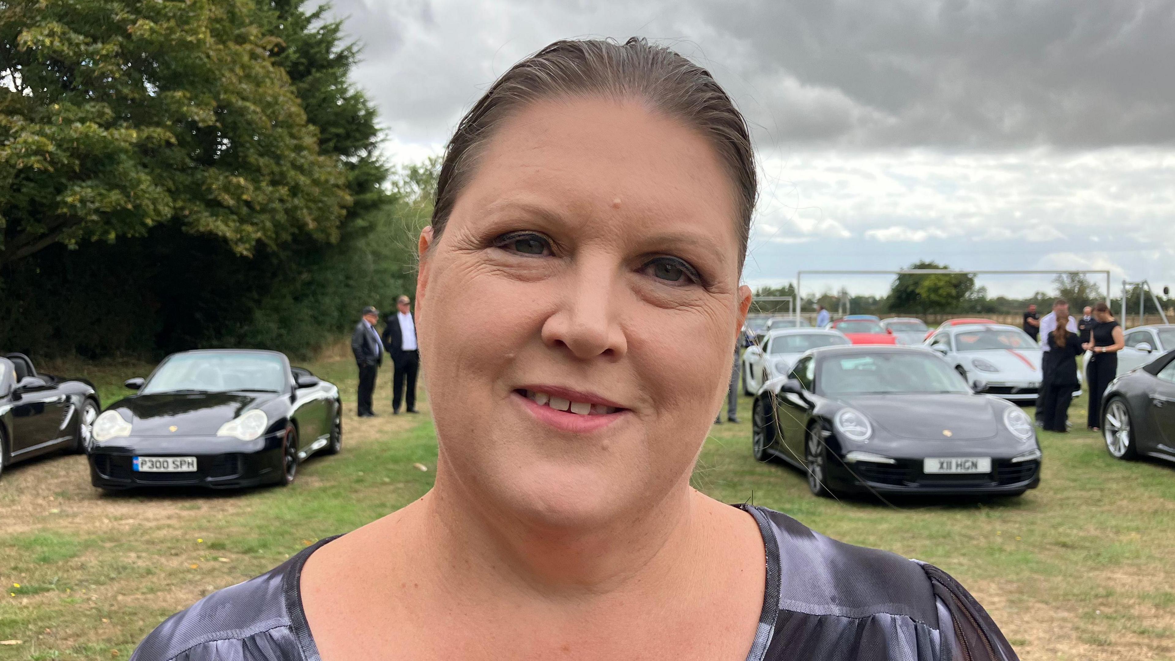 Emma Jackson is looking at the camera with a slight smile. She has dark brown hair in a ponytail and is wearing a black and grey T-shirt. She is standing on a field which has different colour Porsche cars on. 