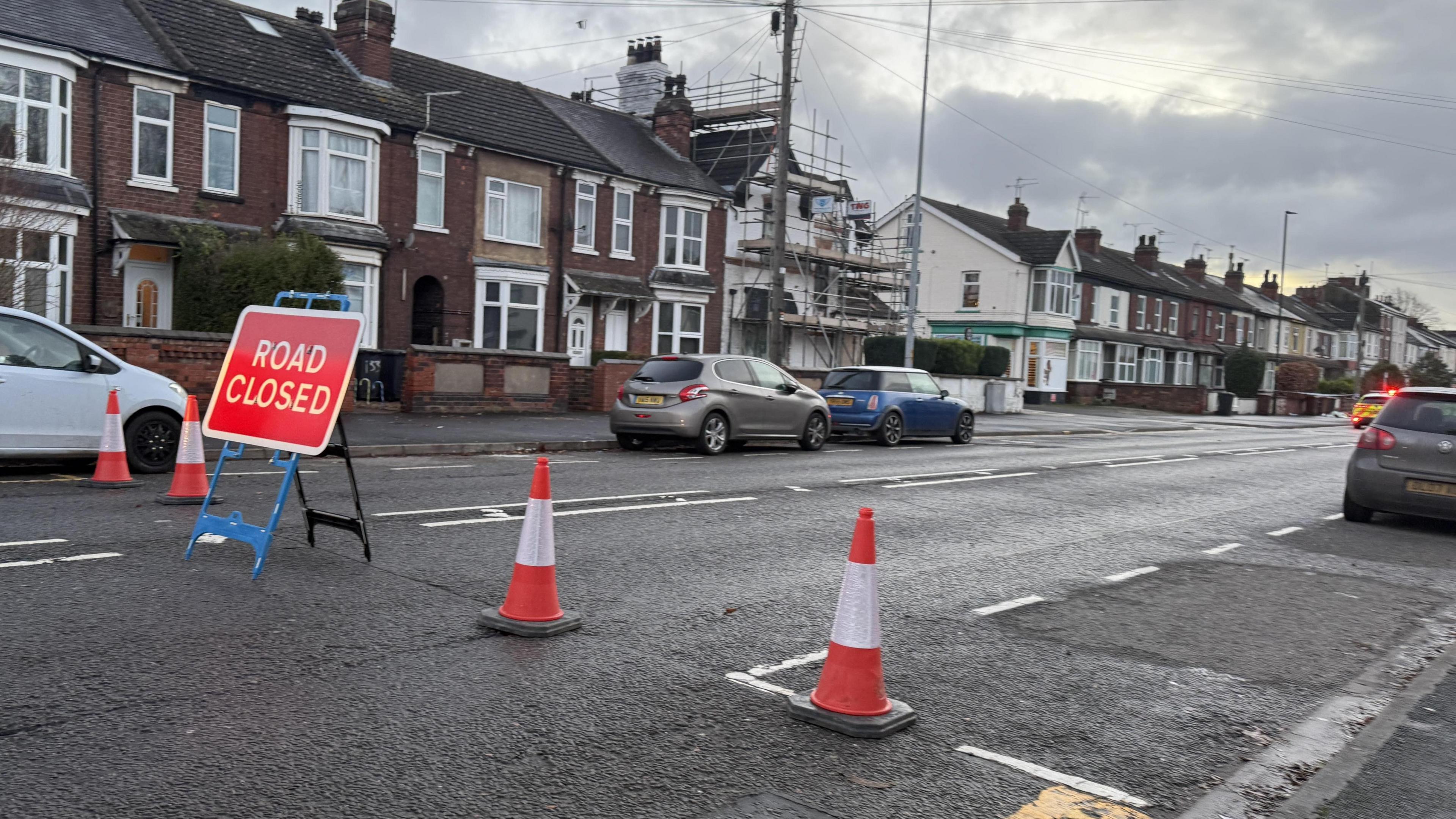 Traffic cones and a "road closed" sign across residential street Carholme Lane. A police car with its lights on can be seen in the background.