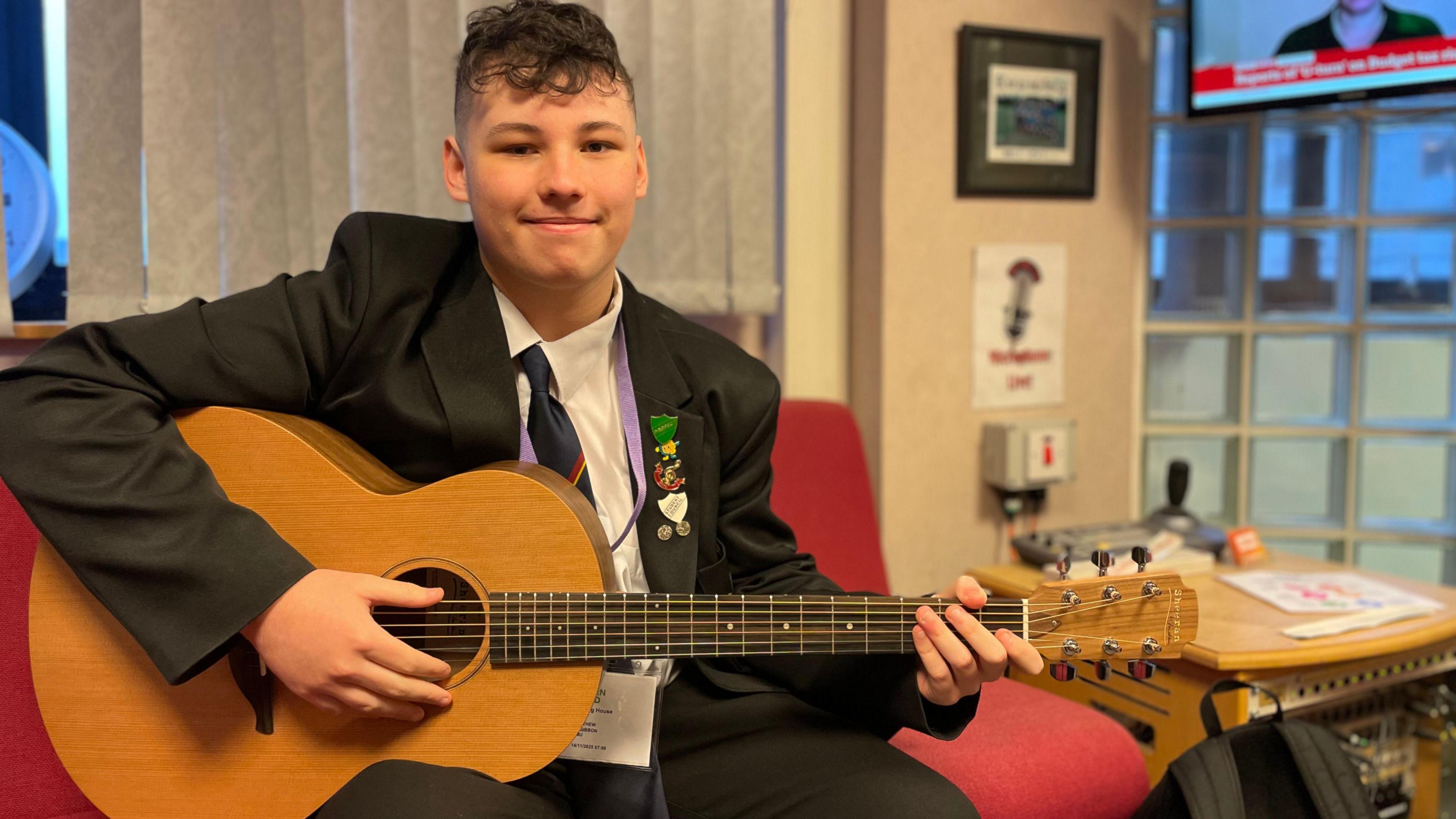 Matthew, a teenage boy, sit on a red sofa holding a light brown acoustic guitar. Smiling comfortably, he is wearing a black school uniform, including a black blazer, white shirt black tie with a red stripe.