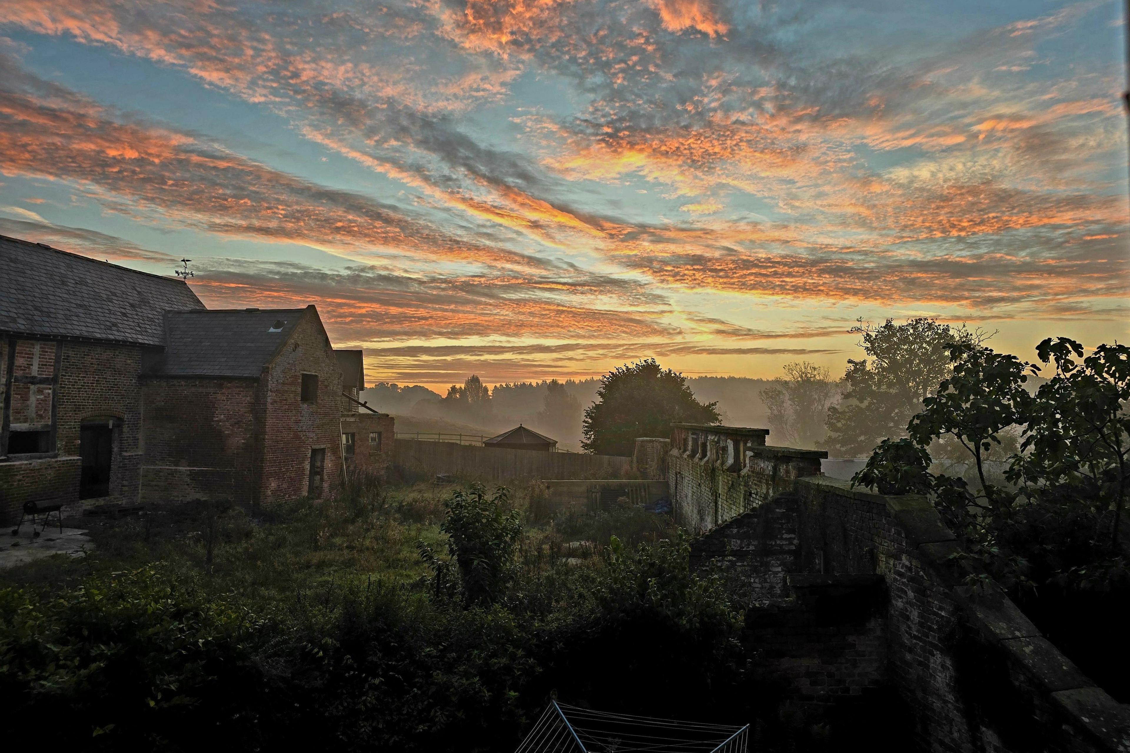 The back of a farmhouse with walled garden under a mainly blue sky with orange-flecked clouds and the sun just over the horizon.