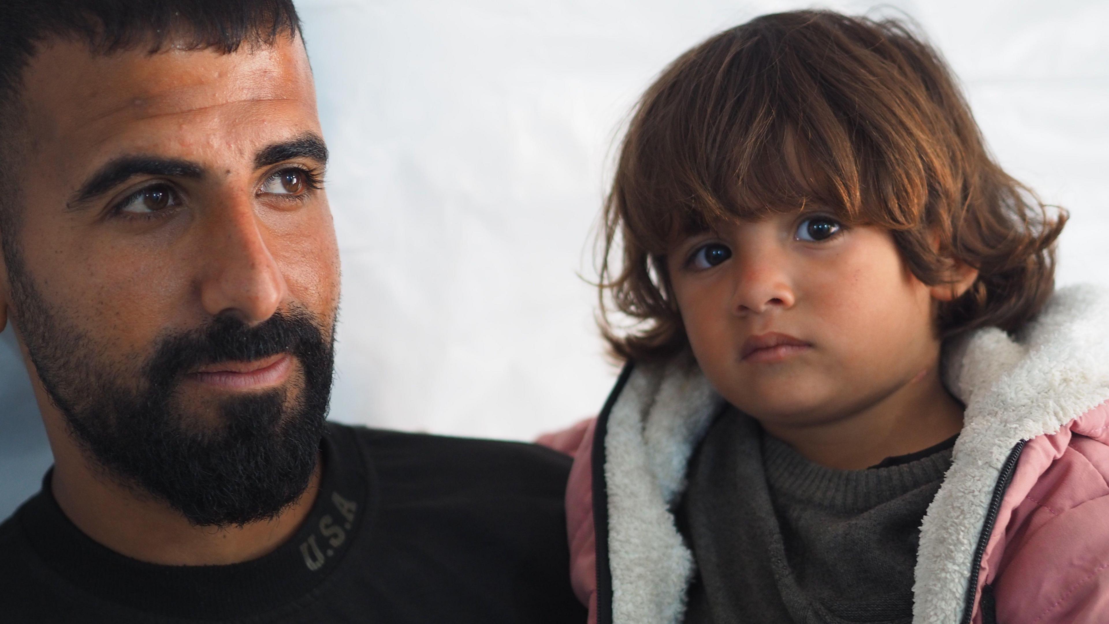 A young child, Razan, is being held in the arms of her father. Her father has a dark beard and a black top on, and looks out of the frame. Razan has a mop of brown hair and large brown eyes and is looking into the camera.