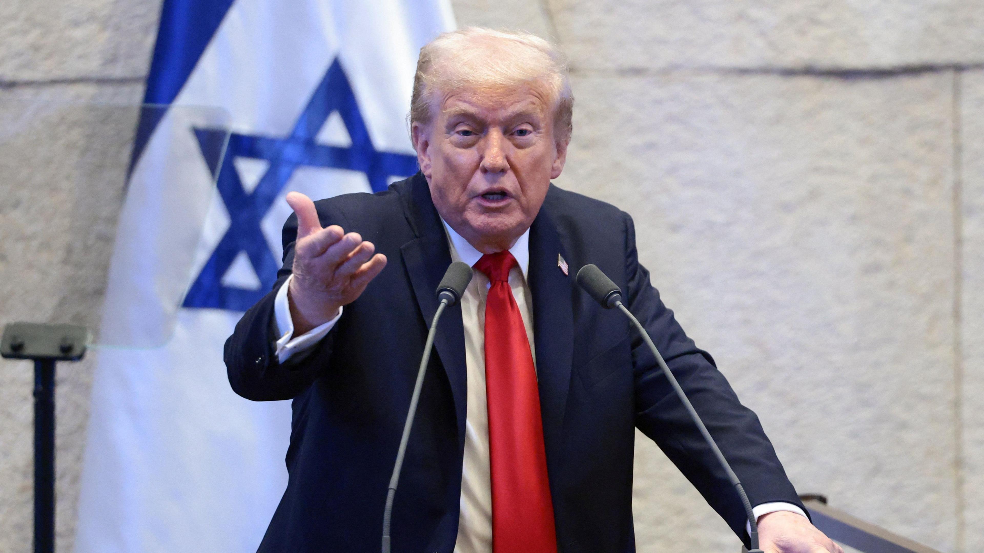 President Trump wears a blue suit, white shirt and red tie. He is making a speech and gesturing with his hand, standing in front of a microphone with the flag of Israel in the background.