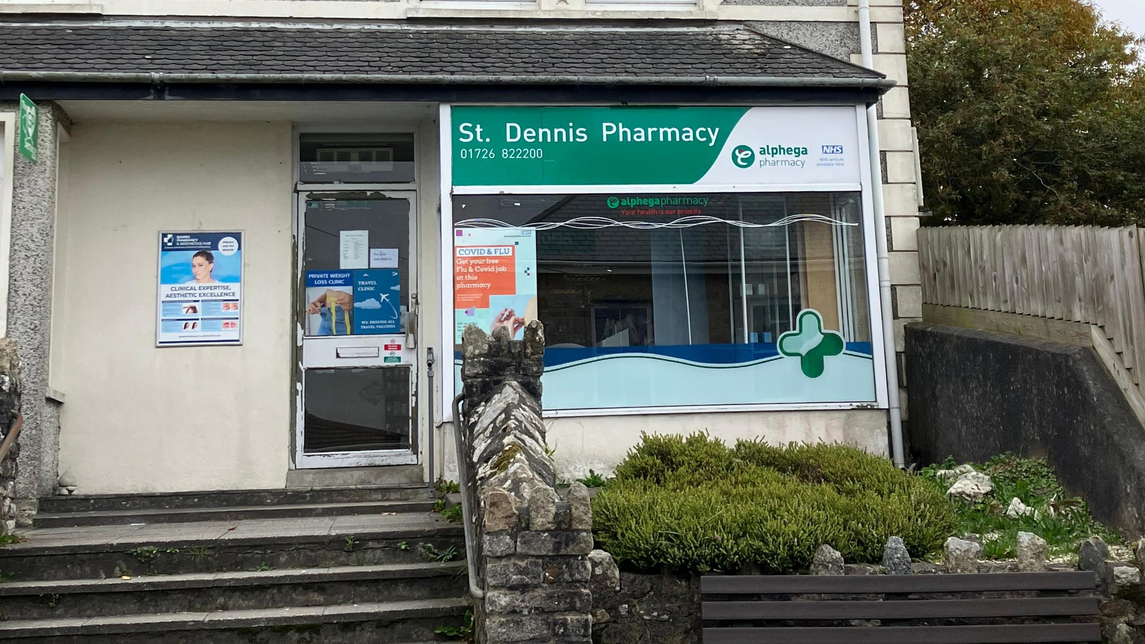 The outside of the St Dennis Pharmacy. Stone steps lead up to a glass front door of a white coloured building with a big front window with a sign with the pharmacy name on top of it. 