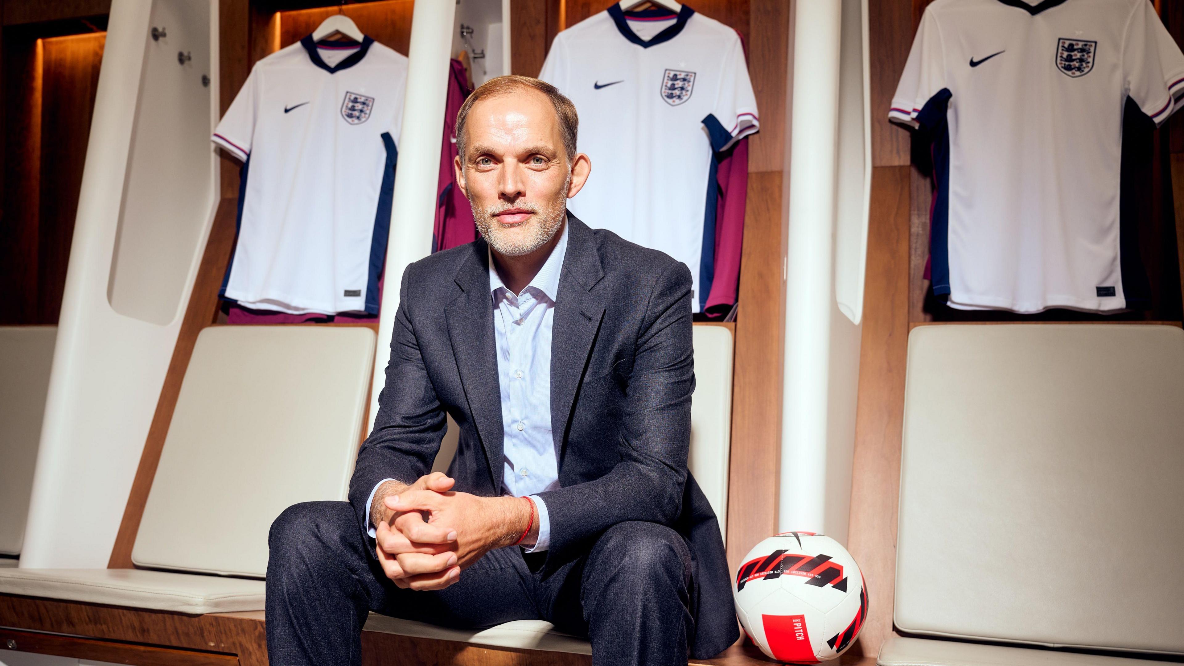 Thomas Tuchel sits down inside a dressing room with England shirts hanging on the wall behind him