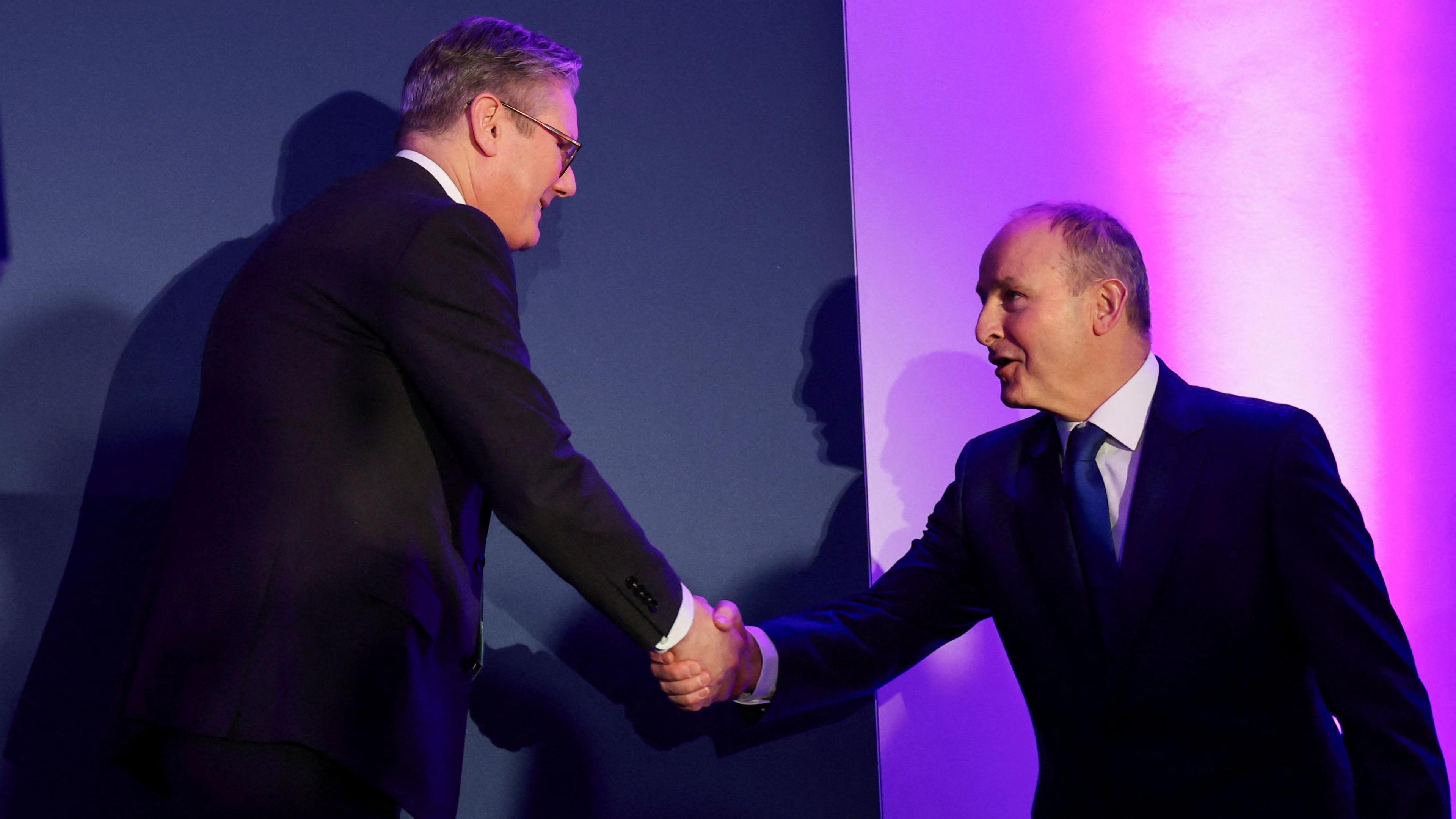 Micheál Martin said Sir Keir Starmer smiling and shaking hands in front of a blue backdrop with purple lighting. They are both wearing suits.