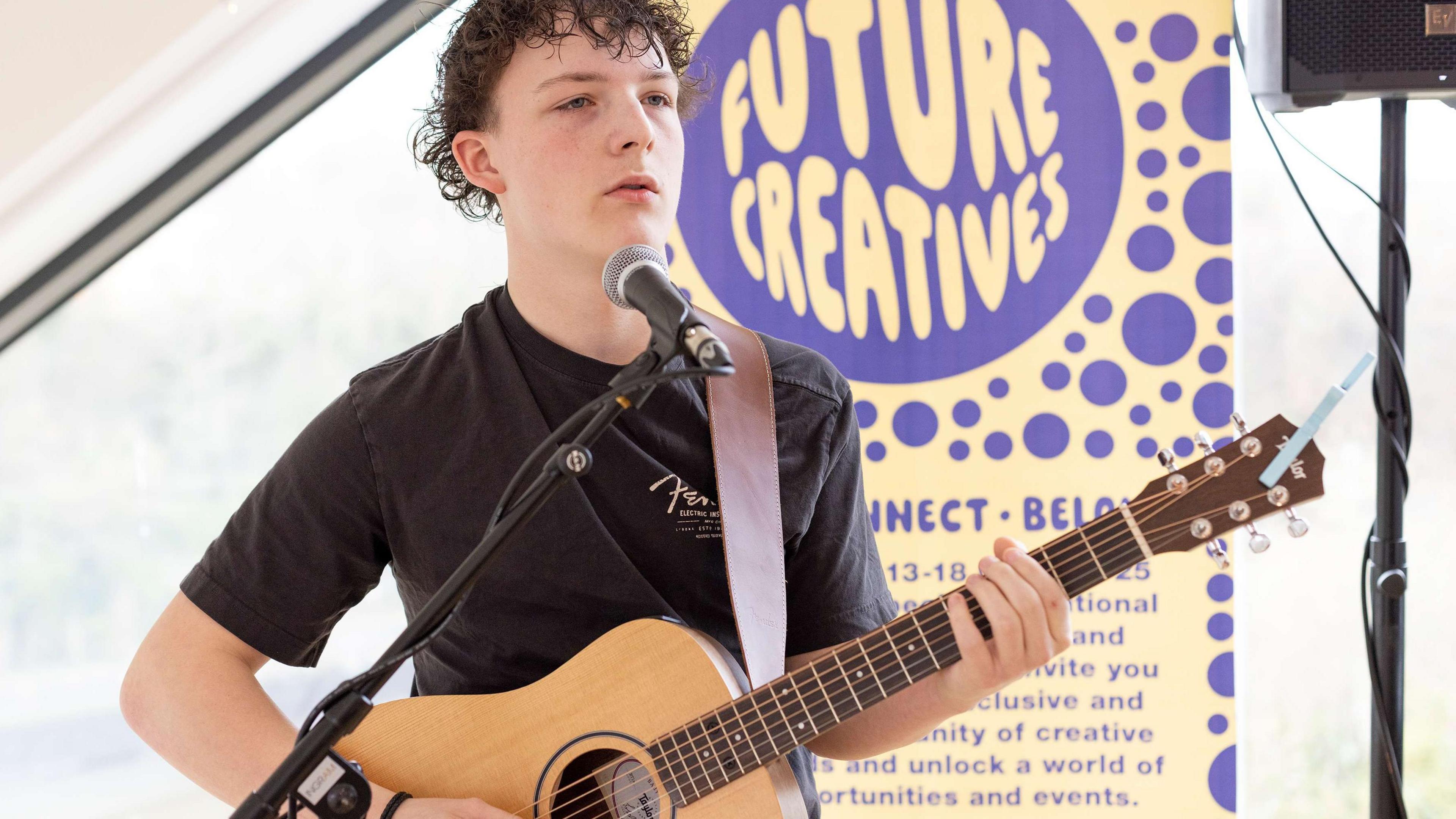 Callum O’Neil, who has wavy dark hair, is wearing dark top and holding a guitar. There is a microphone in front of him and a pull up banner behind him that reads 'Future Creatives'.