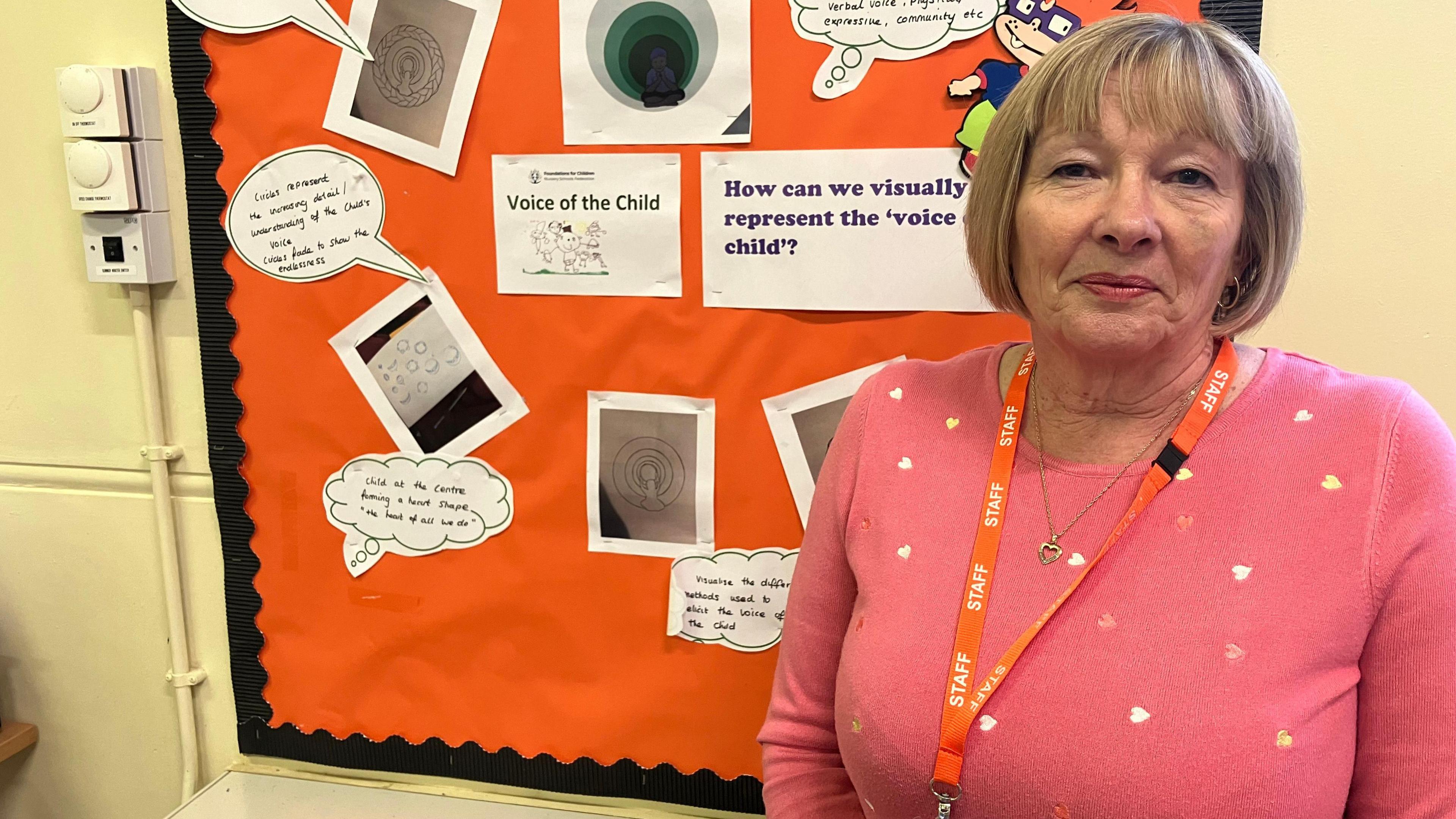 A woman in a pink jumper with hearts on, wearing a orange lanyard, stands in front of an orange board