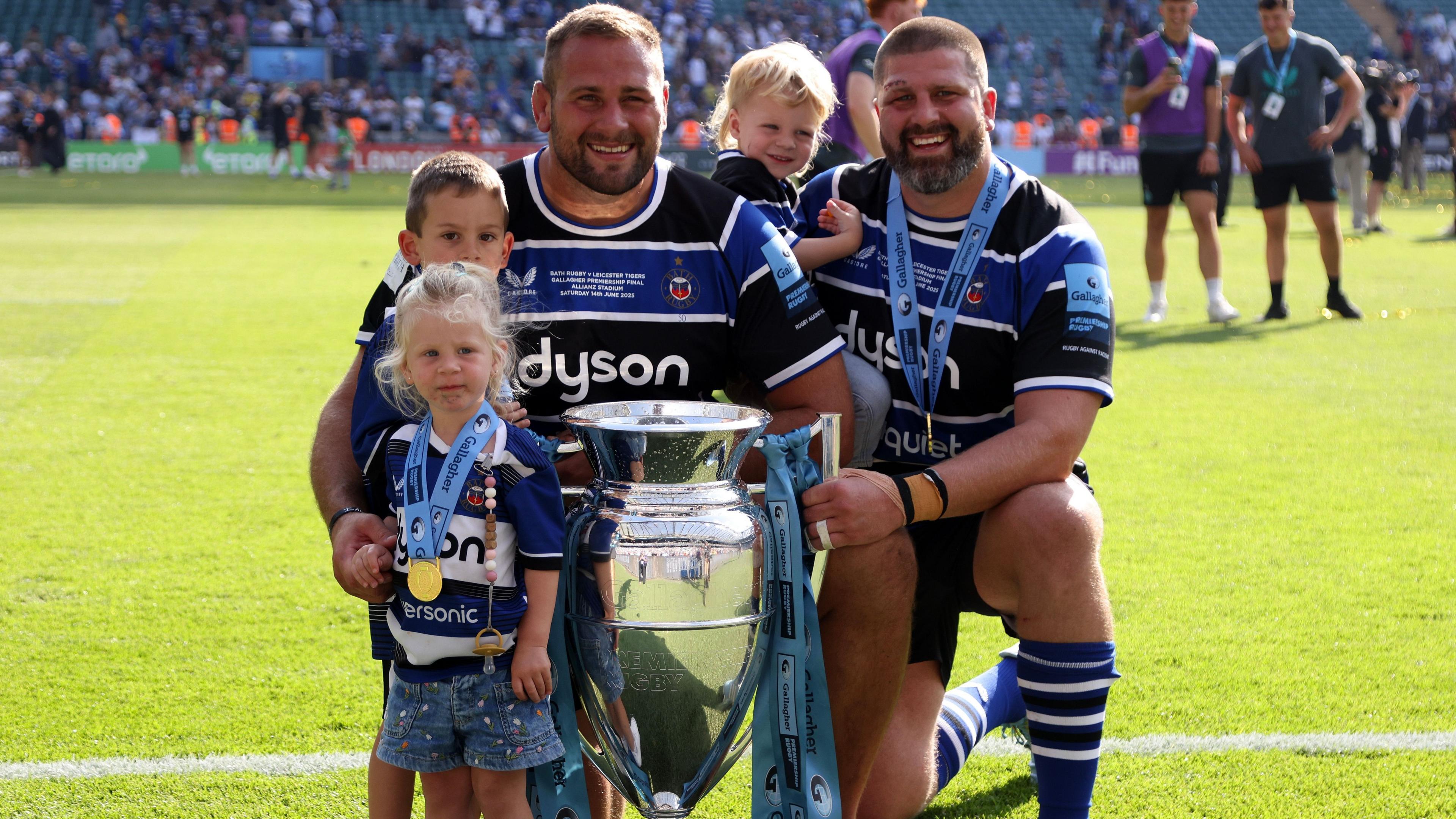 Thomas du Toit and Francois van Wyk kneel on Twickeham turf holding large silver Premiership rugby trophy, surrounded by three young children