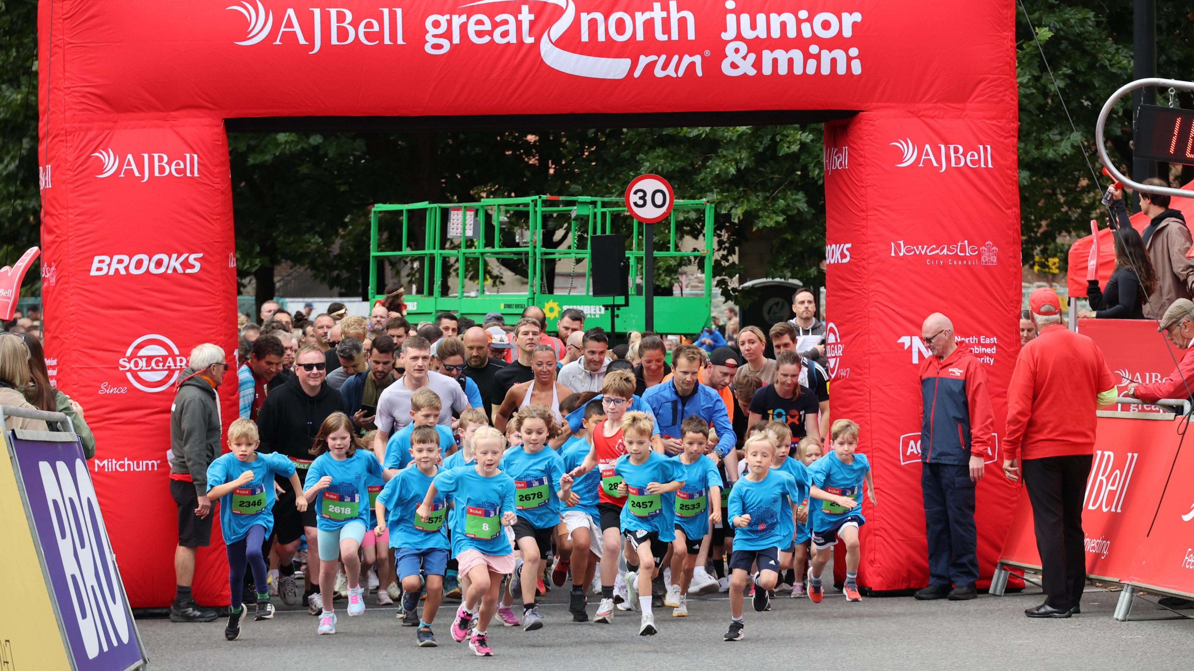 Dozens of small children setting off on foot in a race. They are all wearing blue T-shirts and green signs on their fronts with their race numbers. They are at the starting line of the race which is under a giant red arch that says "AJ Bell Great North Run". Stewards and adults with stand in the background