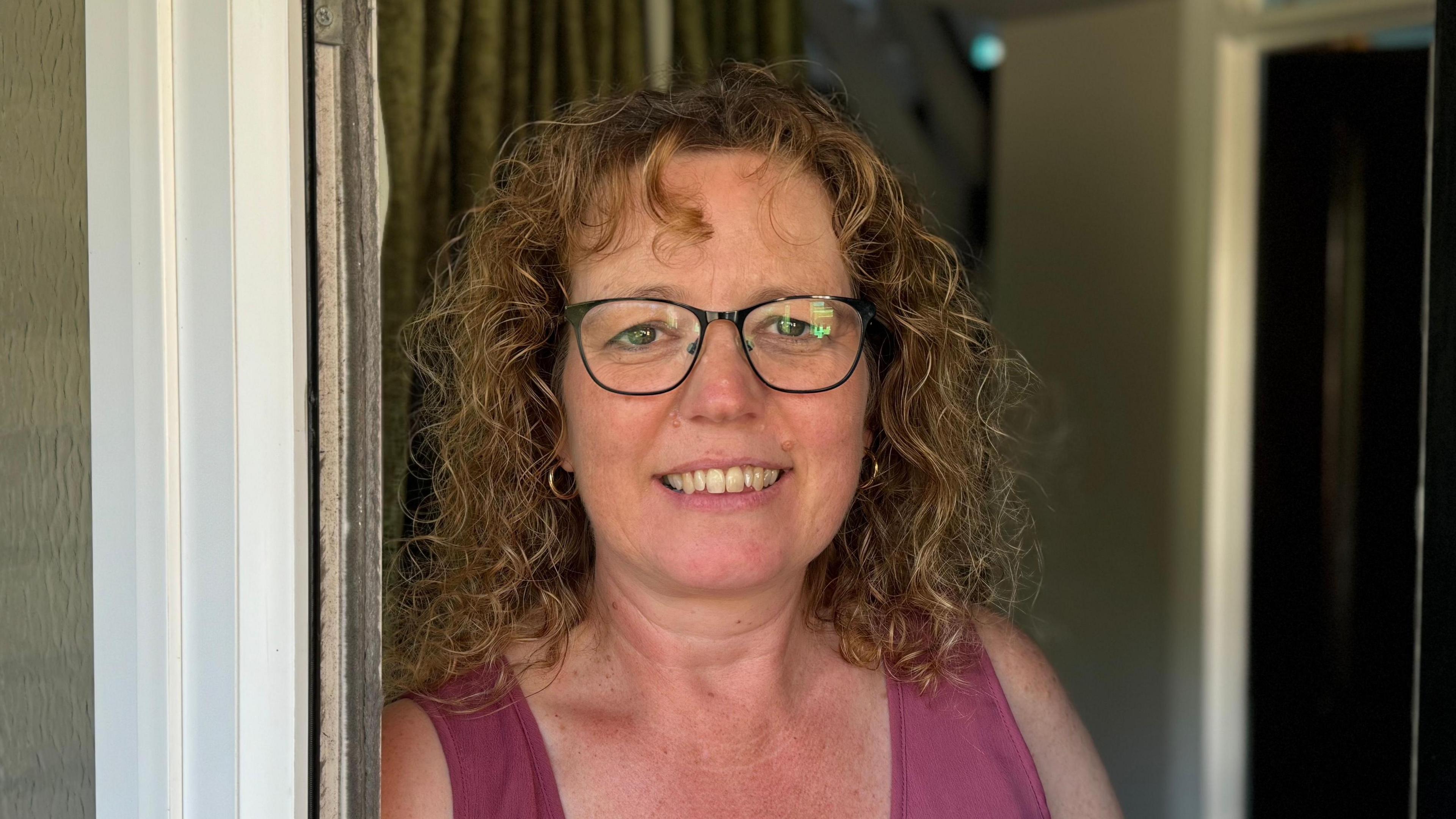 A woman with curly hair and glasses, wearing a pink vest top, smiling into the camera