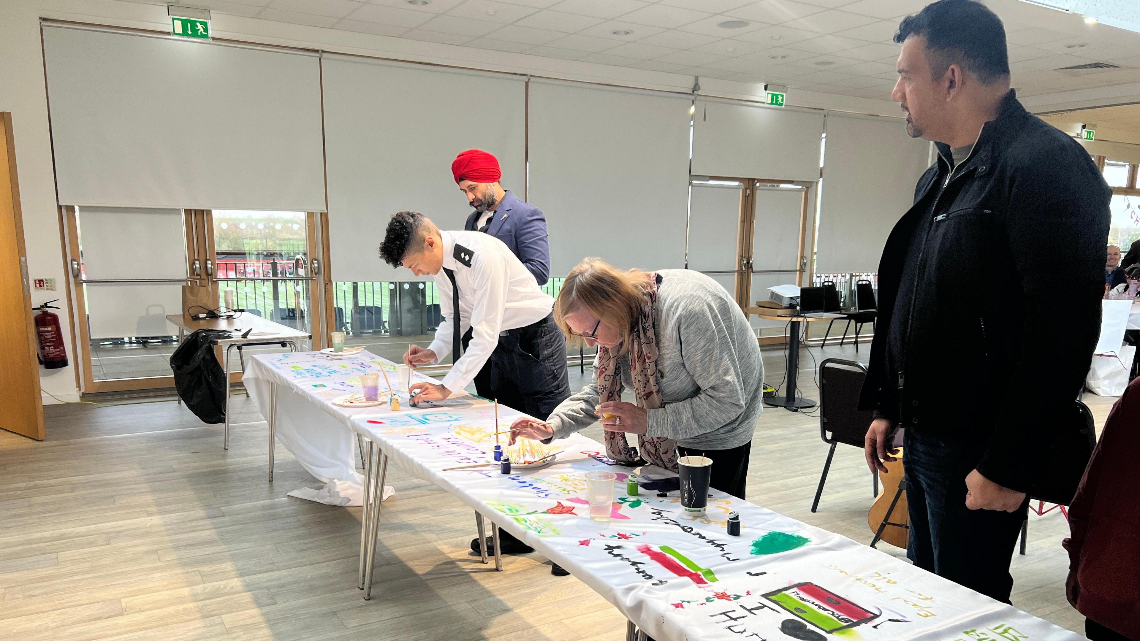 A man in red turban, a police officer in uniform, a woman in a grey sweater and flowery scarf painting on a long white piece of silk. Next to them is a man dressed in black.