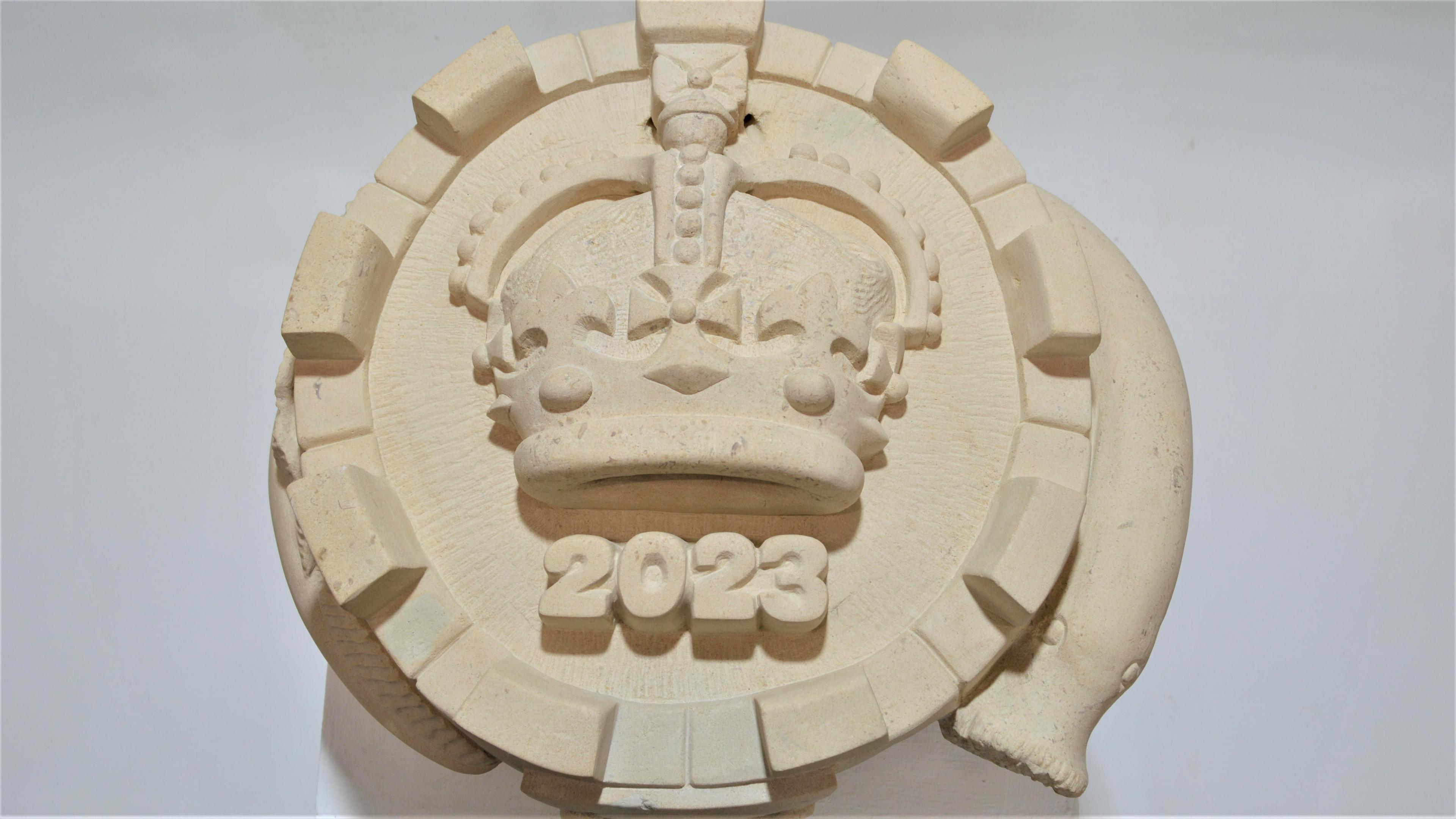 A circular piece of stone with a monarch's crown in the middle, 2023 written underneath it and a lamprey snaking around its sides.