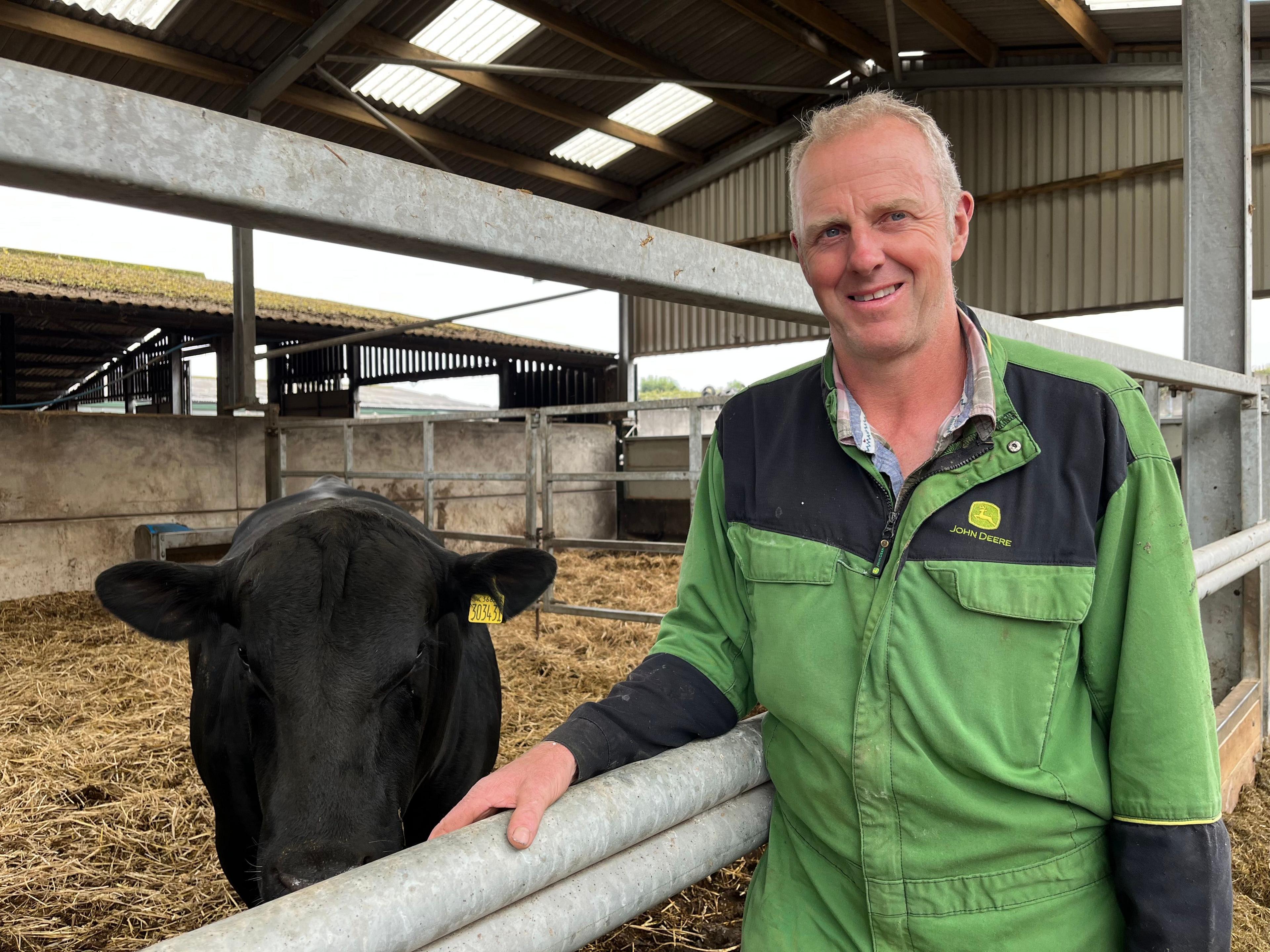 Andrew Jones is wearing green overalls. He is standing in front of a cow in a barn.