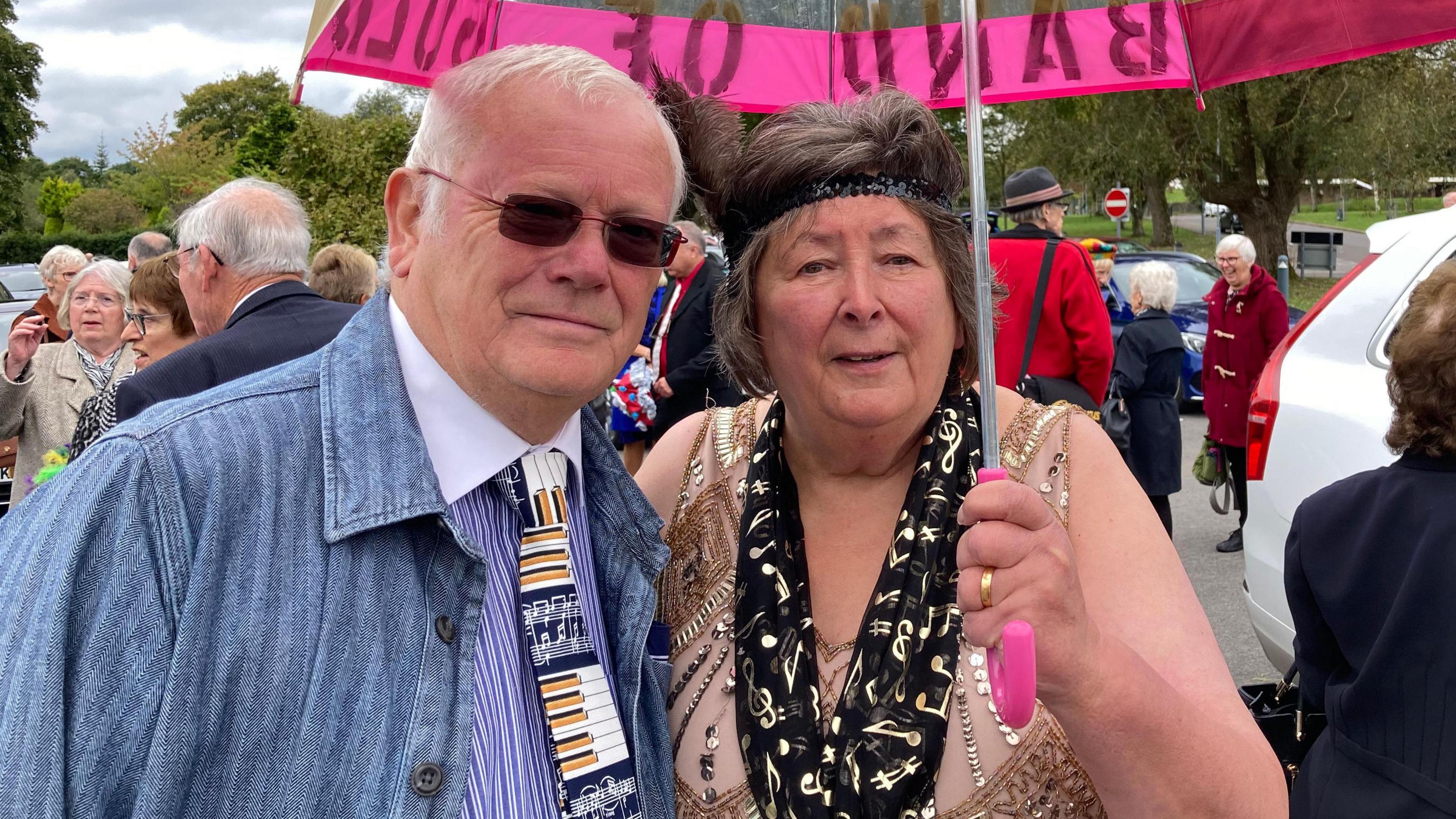 Howard and Carol smiling at the camera underneath a pink umberalla at the funeral - Carol is wearing a 1920s style headpiece and gold dress