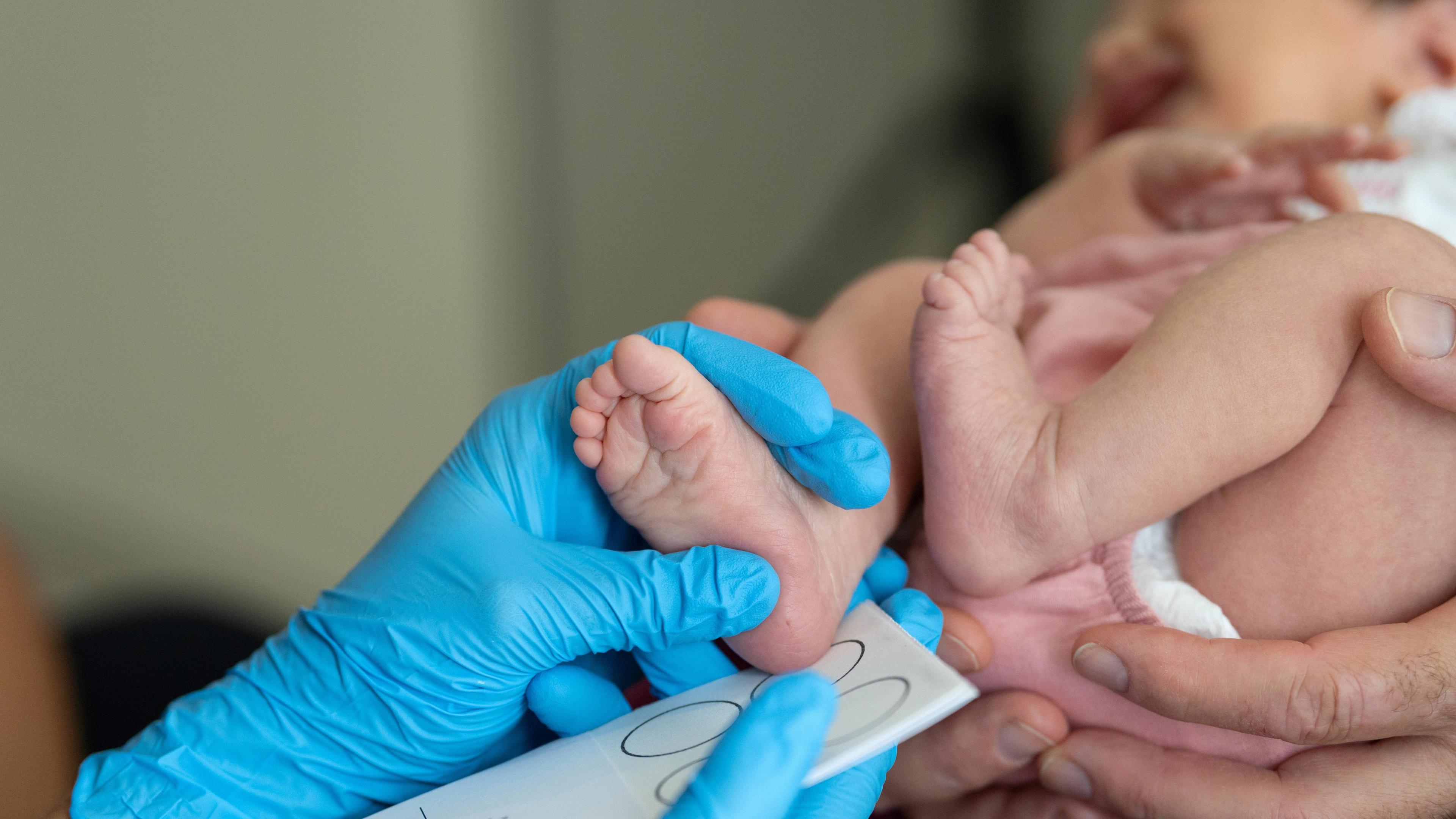 The hand of a doctor or nurse holding a new-born baby's foot and measuring it against circles on paper. They are wearing blue gloves. The baby is wearing pink. Hands are holding the baby up.