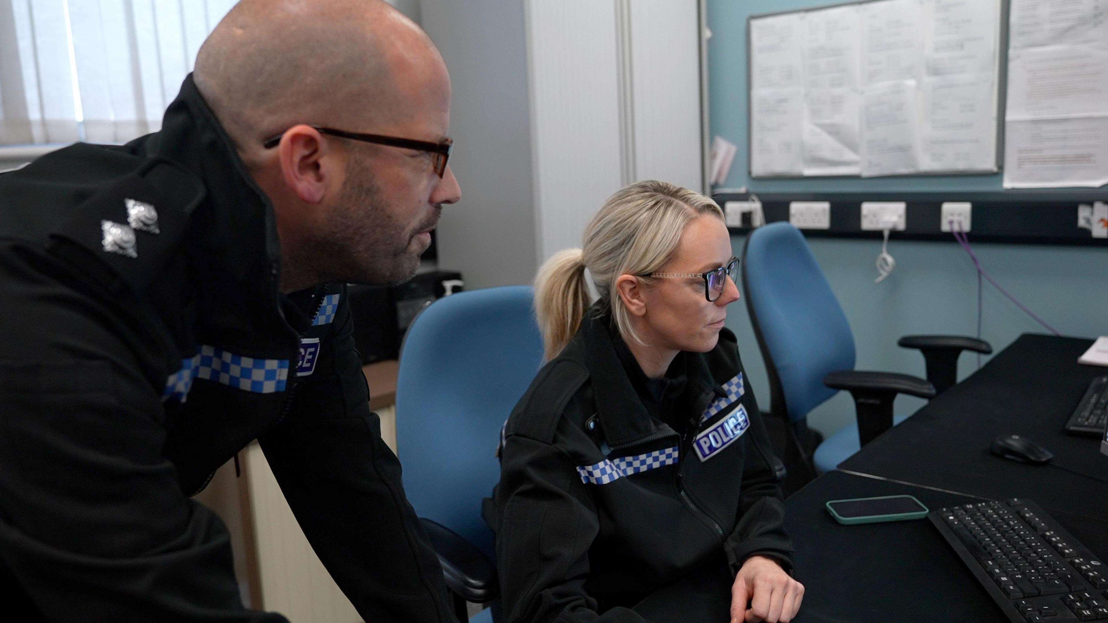 A male police officer stands next to a female officer who is sat at a desk looking at a computer