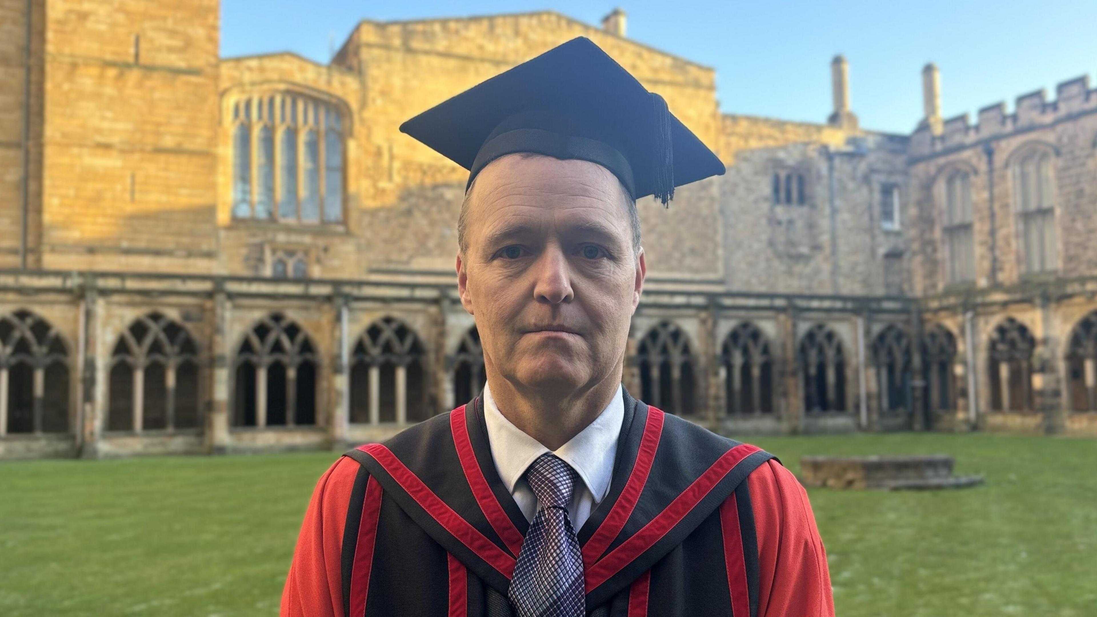 A man wearing a red and black cloak with a black graduation hat, standing in front of a cathedral. 