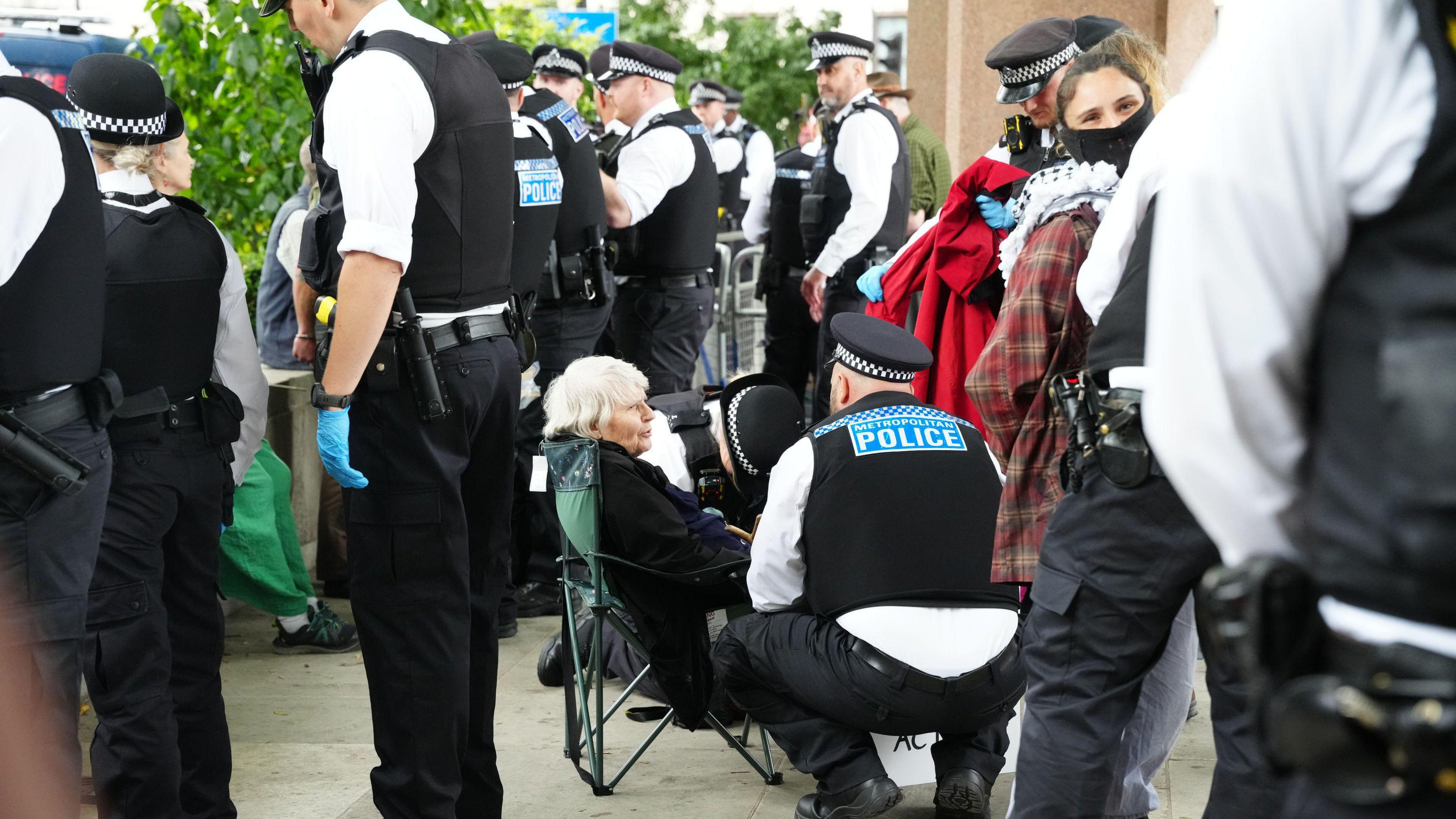 Reverend Sue Parfitt sitting in a green camping chair, speaking wit an officer who is crouched down beside her. They are surrounded by more police officers wearing black and white uniforms and stab proof vests.