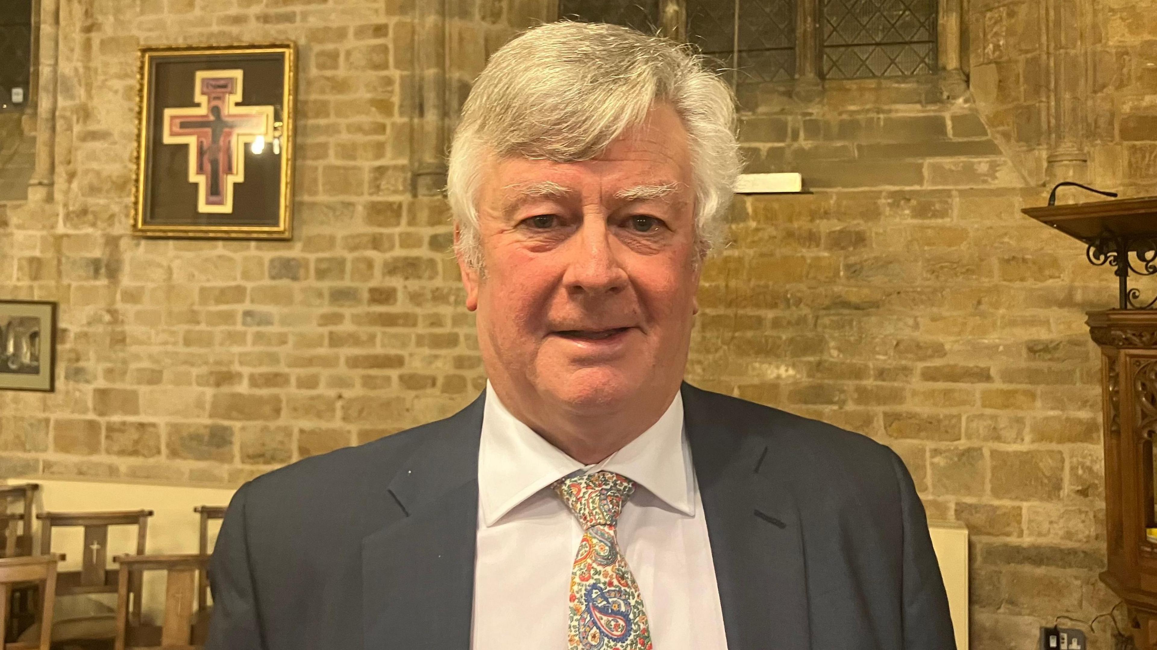 Graham Ferrie, who has short, white hair and is wearing a black blazer, white shirt and a multi-coloured tie. The picture has been taken in a church. Chairs, a brick wall and part of the pulpit are in the background.