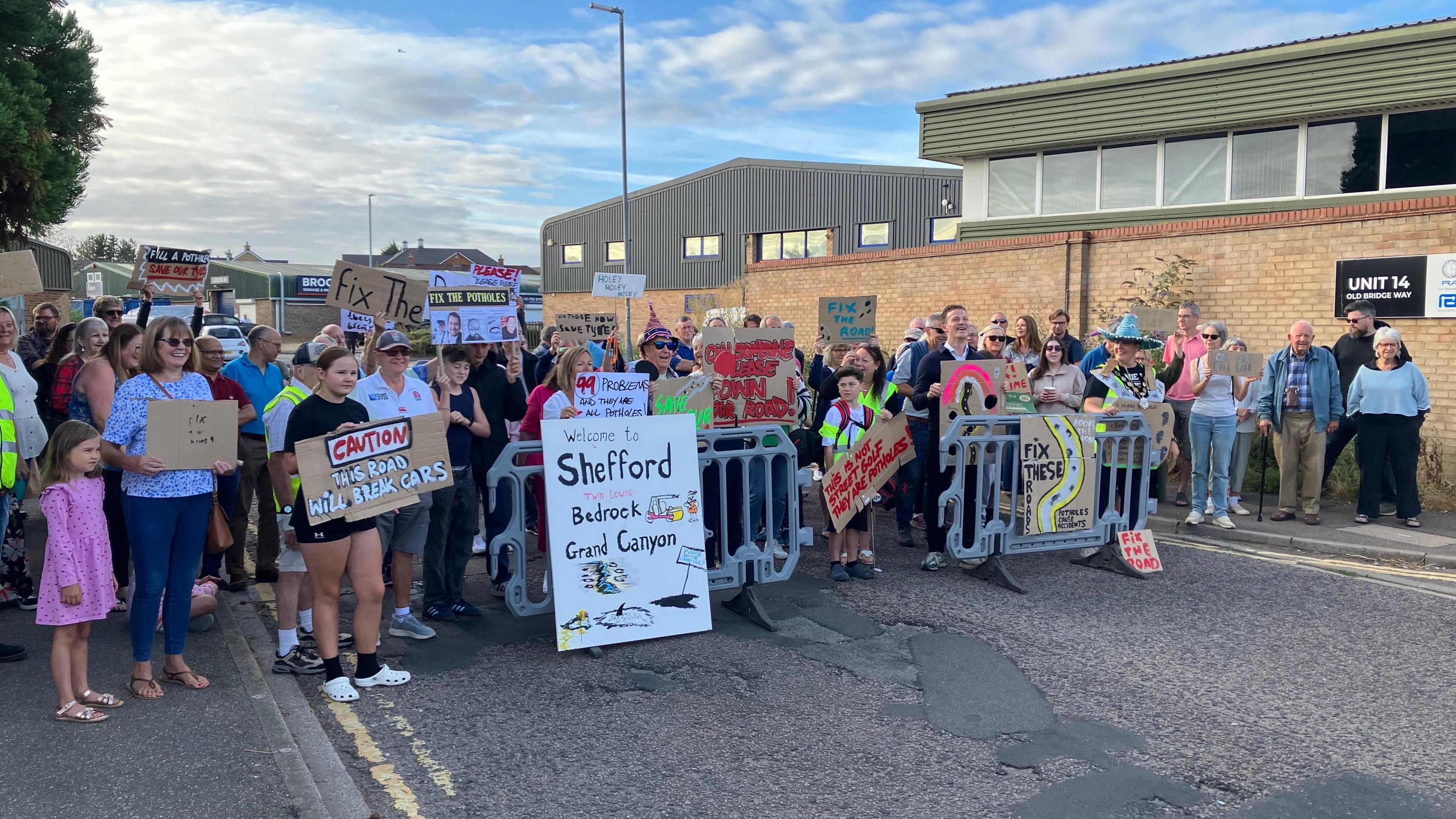 A picture of protestors who have blocked a section of road in Shefford in Bedfordshire. They are holding various signs to campaign for potholes to be filled in on the road. There are men, women and children of a variety of ages. The sky is blue behind them and most are stood on the road.