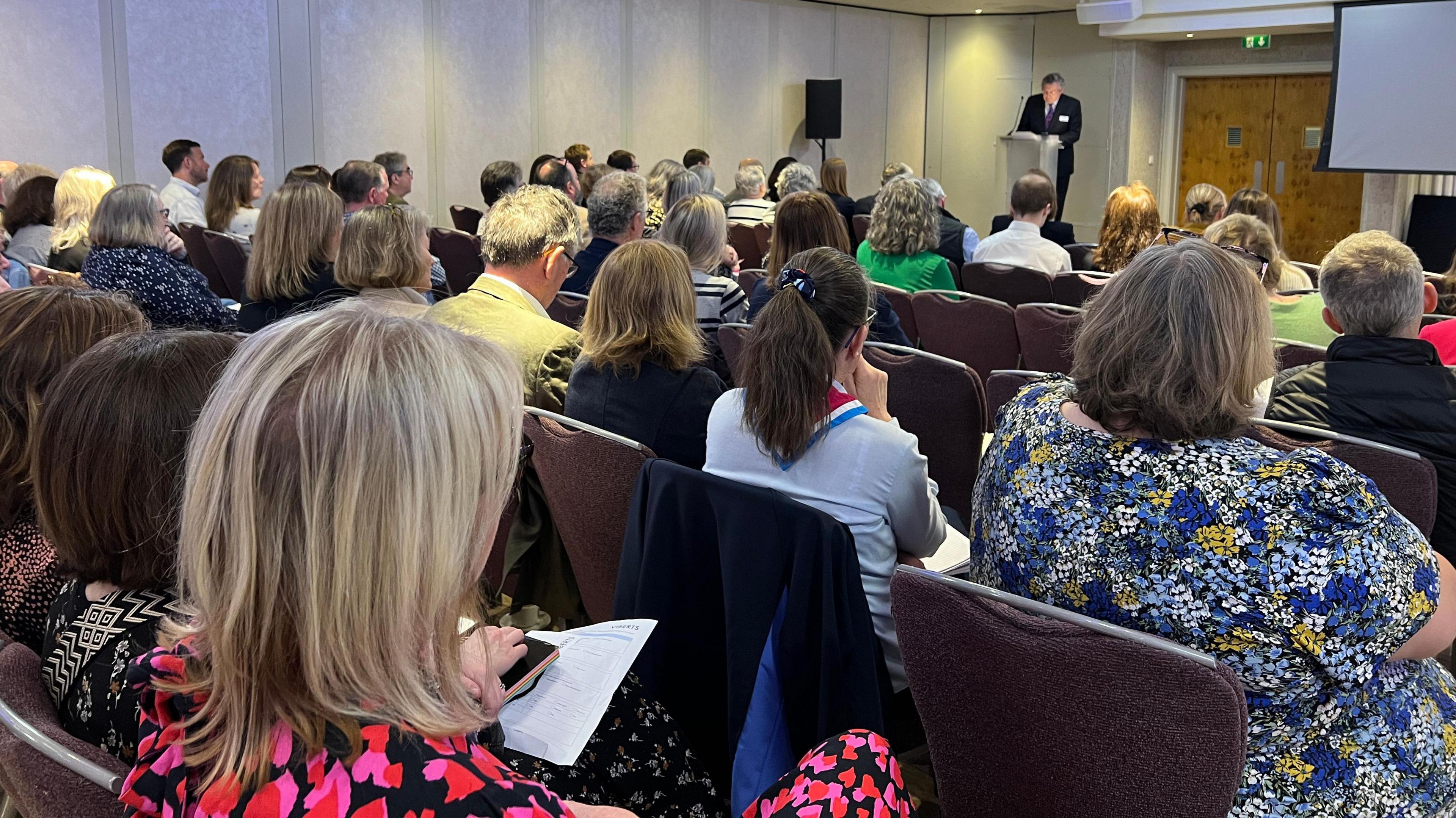 A crowd watches John Mills give his speech in front of a podium at the conference