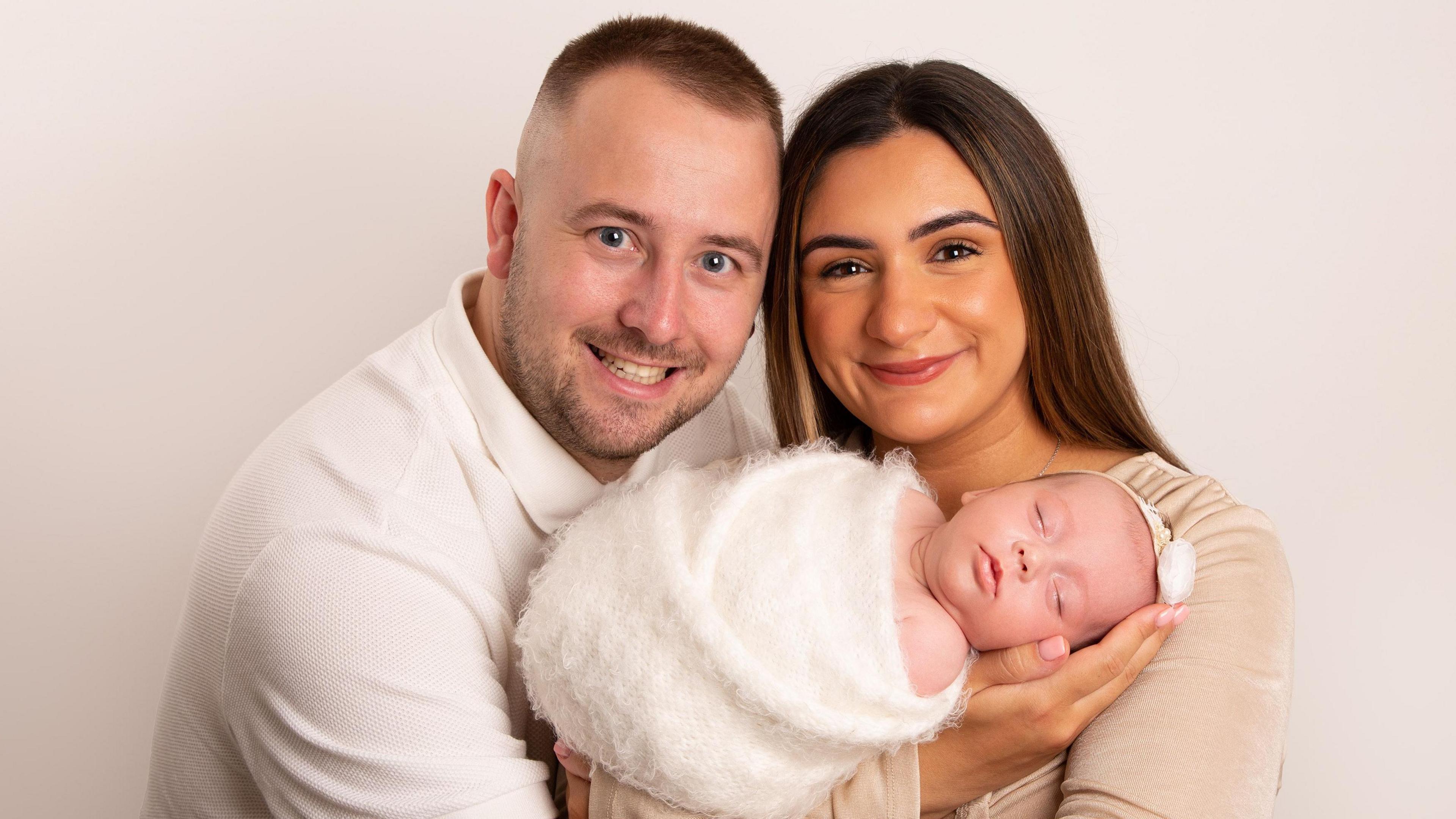 David and Aisha McCracken with their baby daughter Sophie in a studio shot. The baby is sleeping, wrapped in a furry, white blanket. Both parents are smiling at the camera. 