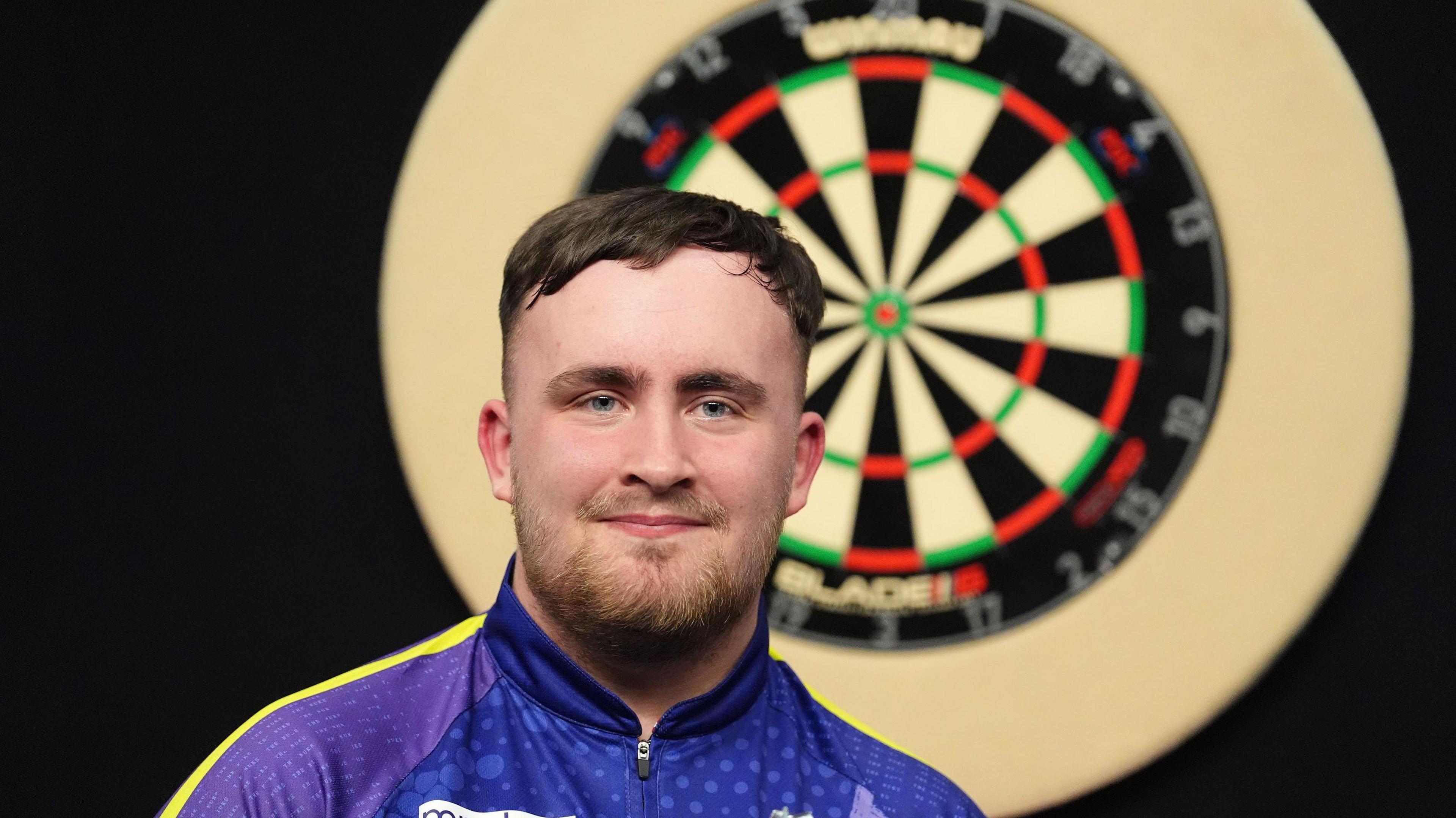 Head-and-shoulders shot of dart player Luke Littler wearing a purple zip-up sports shirt, with a dart board in the background