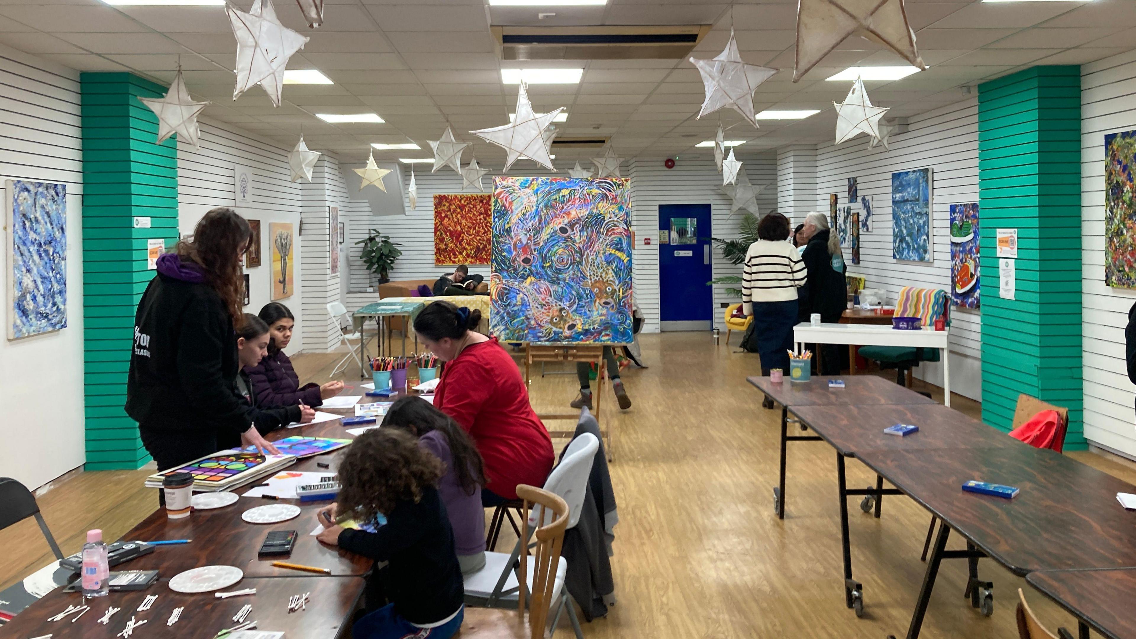 A view of inside of Crewe Makes, with children and adults working on art pieces. Paper stars are hanging from the ceiling, and paintings can be seen on easels.