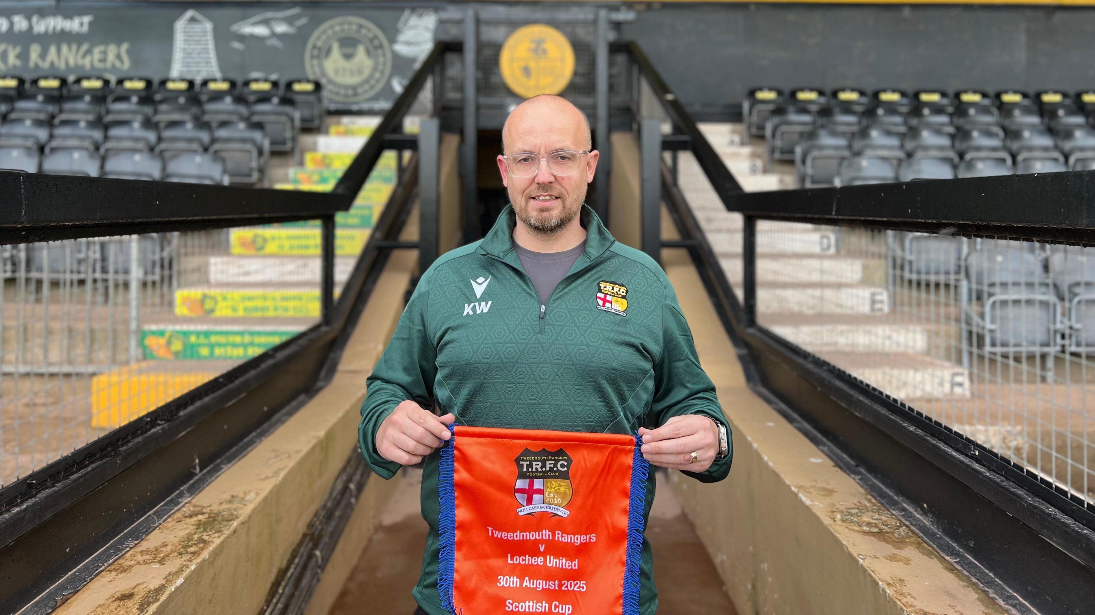 A bald-headed man with glasses and a beard, wearing a green track top, and holding a pendant, stands in front of a football stand