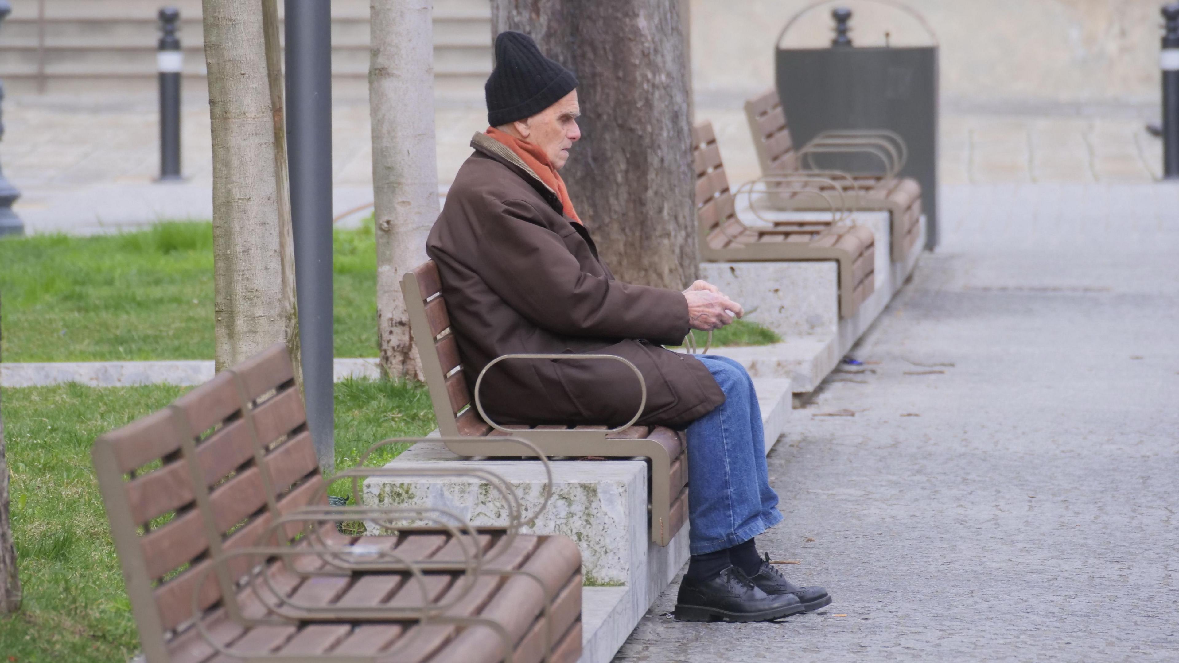 A man is sitting on a bench to the side of a path with grass behind him. He wears a black woolly hat, an orange scarf and brown coat, blue jeans, and black socks and shoes.