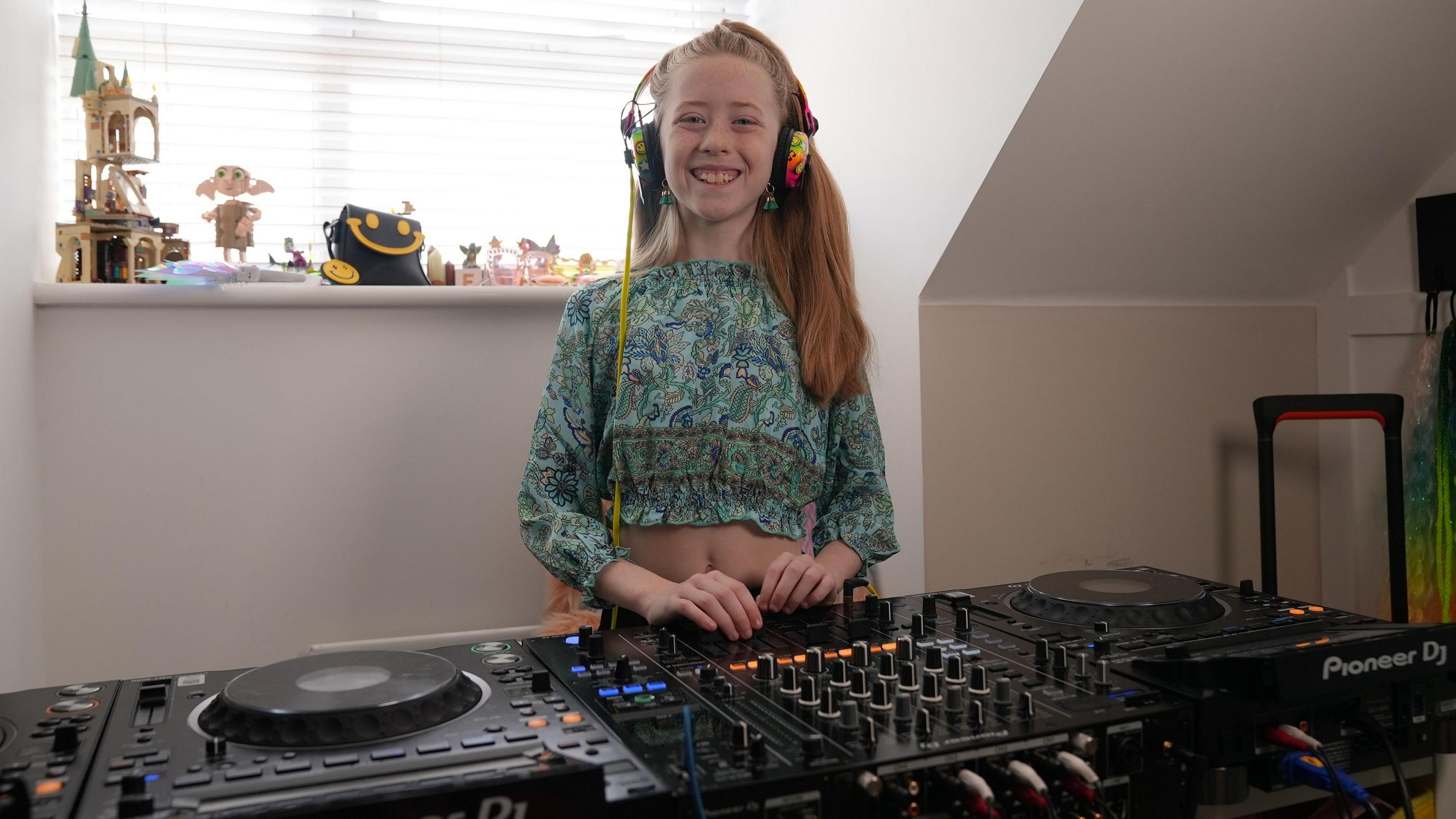A young girl with long blonde hair is in her house smiling and stood behind some large black DJ decks full of dials and lights