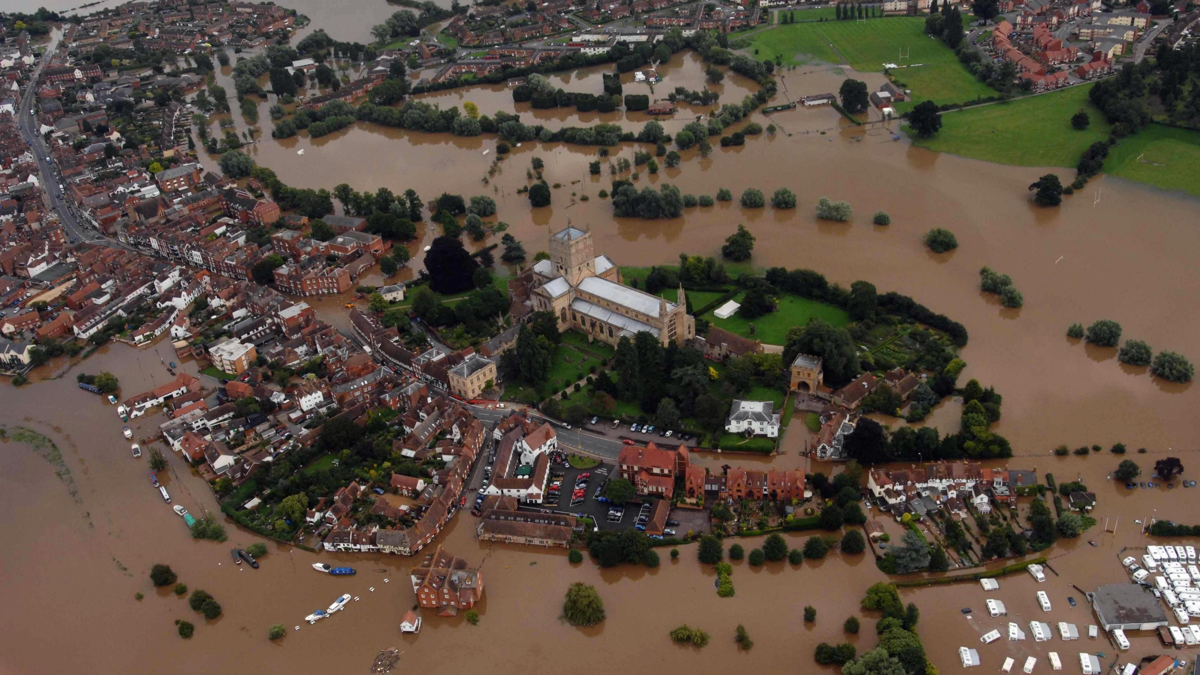 An aerial view showing Tewkesbury during widespread flooding. Brown floodwater are seen on most of the town, with the church one of the few places not flooded