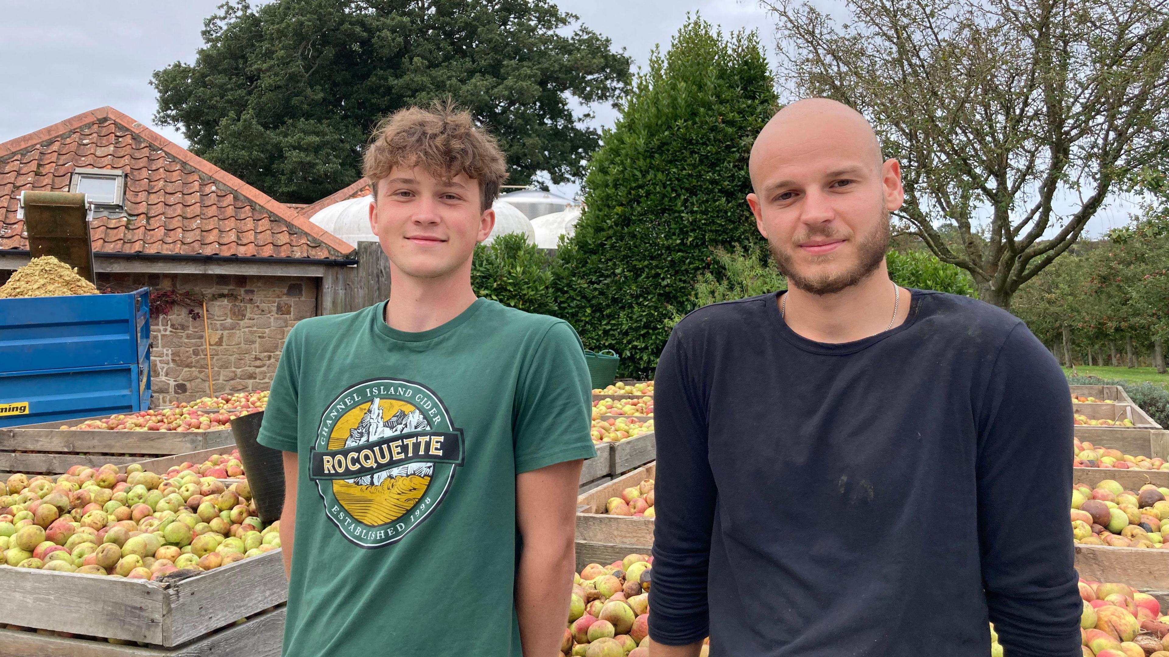 A man in a green and yellow t-shirt standing in front of apples, next to a bald man wearing a blue top