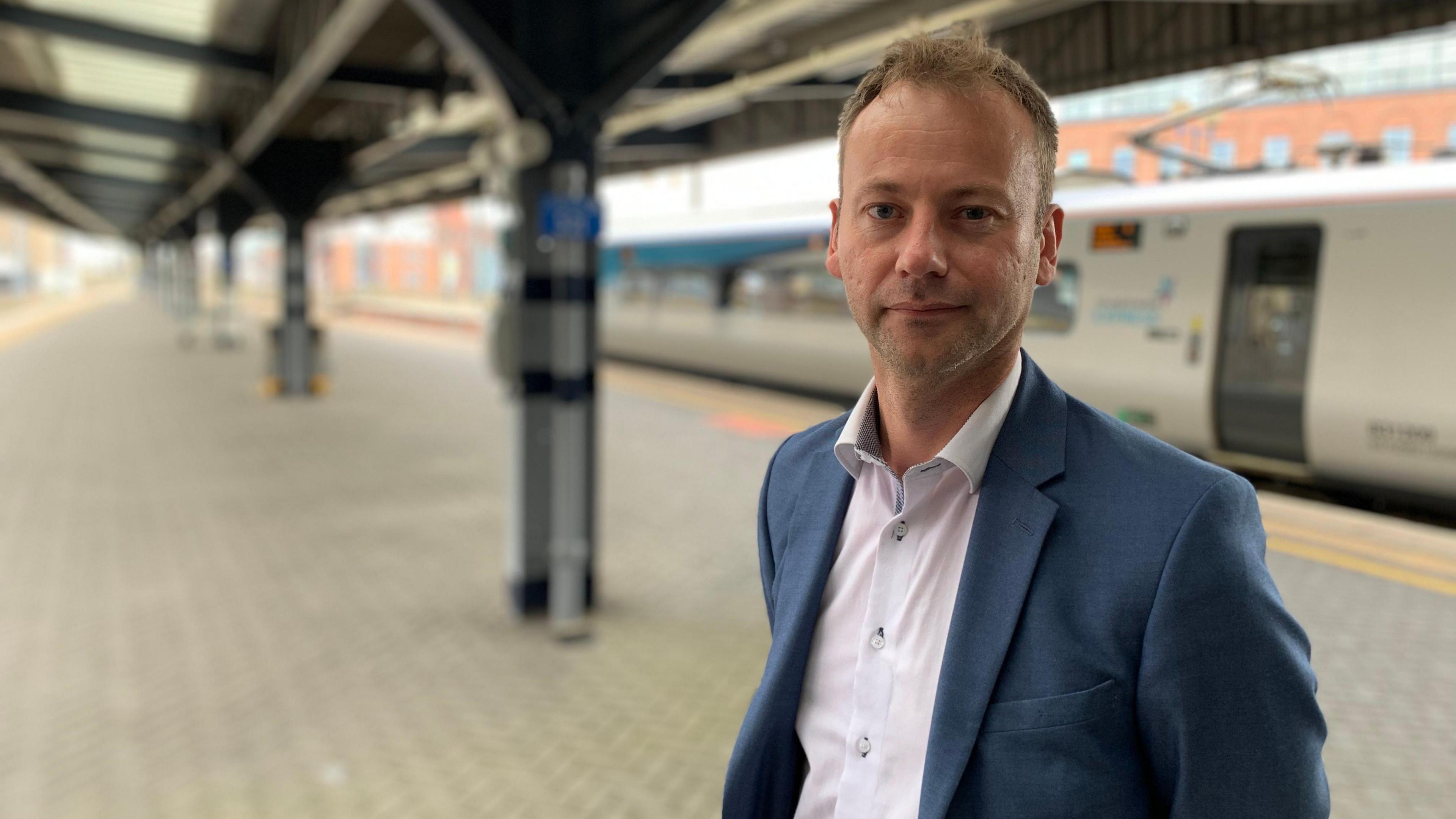 Richard Wearmouth looking into the camera with a straight face. He has brown, short hair and is wearing a light shirt with blue suit jacket. He is standing on a train platform and a train is on the platform behind him.