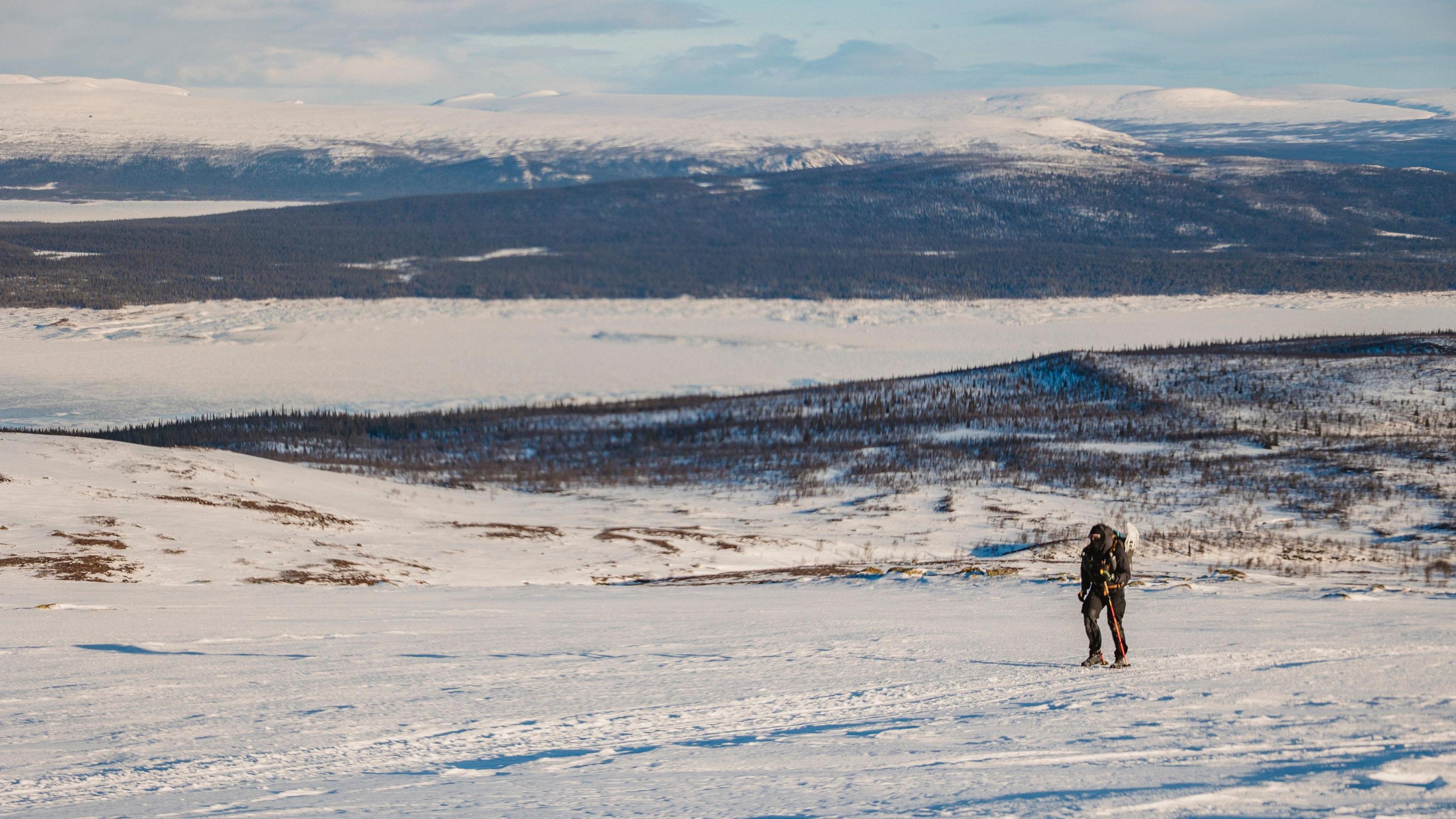 A picture of the tundra and Marc Rhodes climbing up a steep hill. Much of the scene is covered in snow, other than mountains which appear black and have not been covered in it.