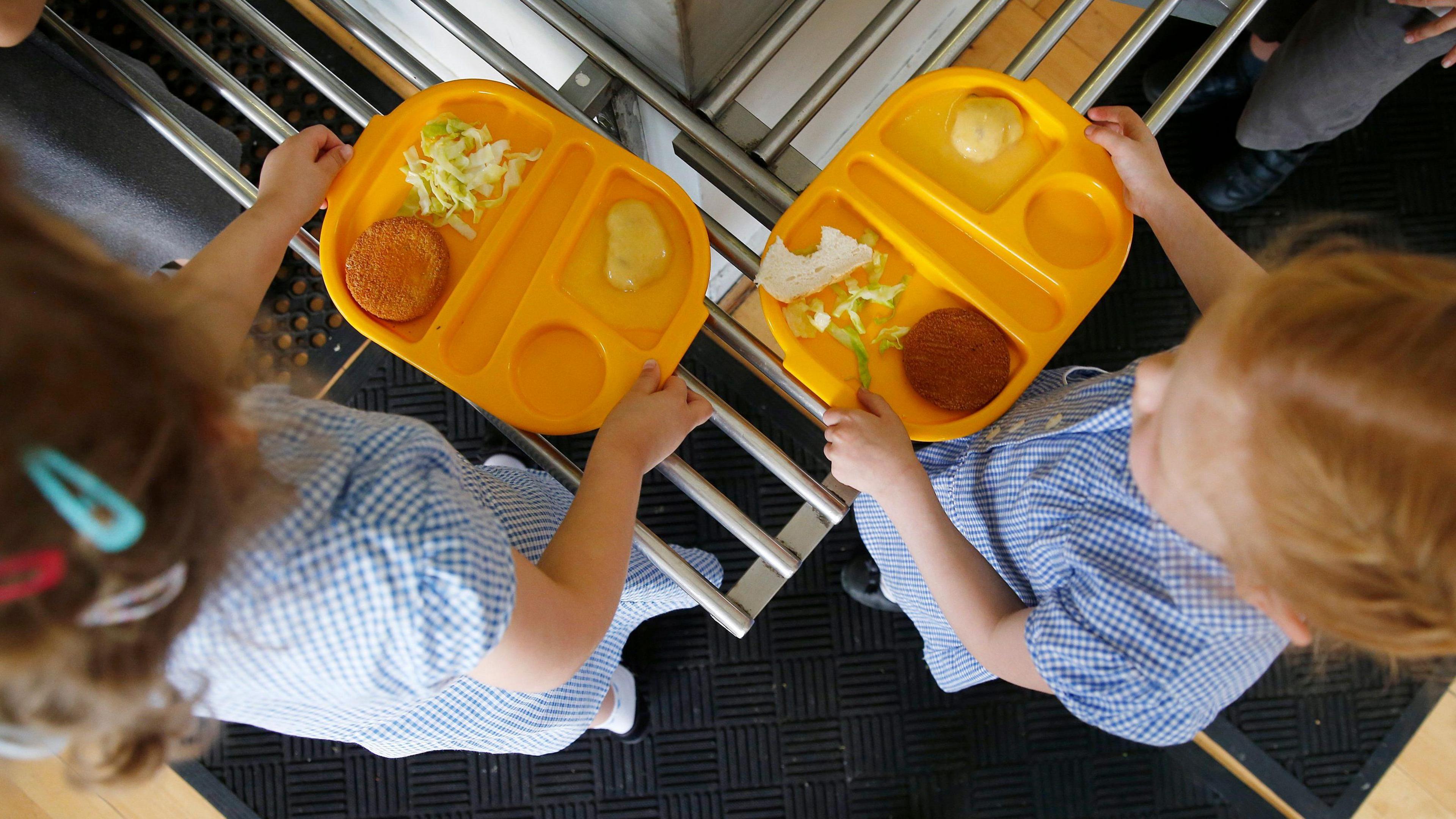 Two young children, photographed from above, both wearing a blue school uniform and hold bright yellow plastic trays. On the trays are some shredded lettuce and some other foods. 