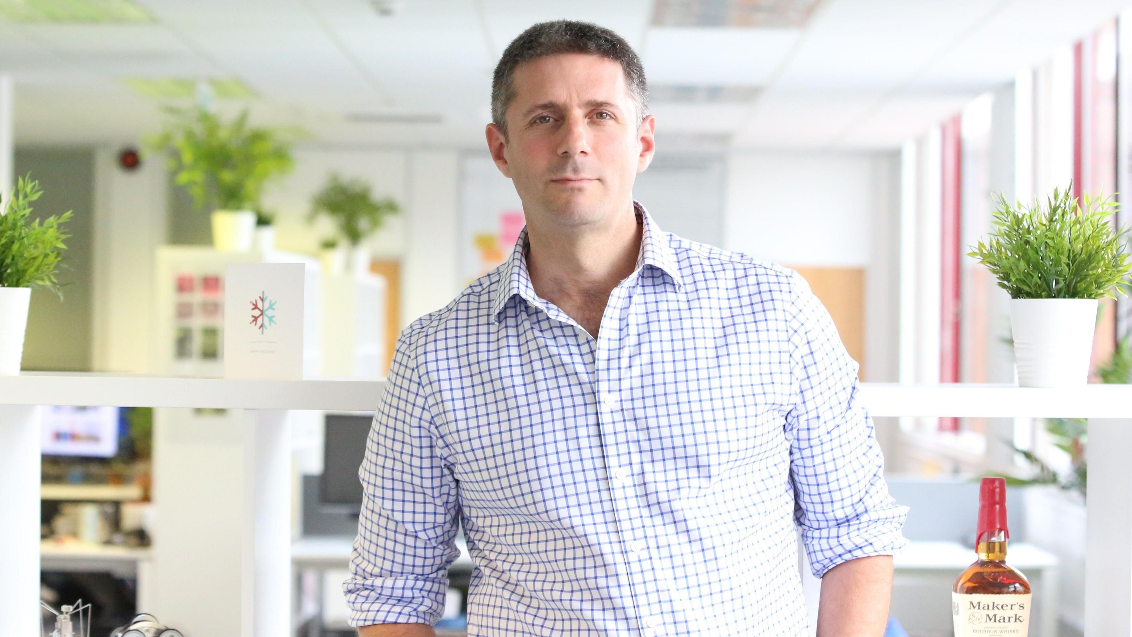 Richard Broadbent standing in an office wearing a white and blue checked shirt