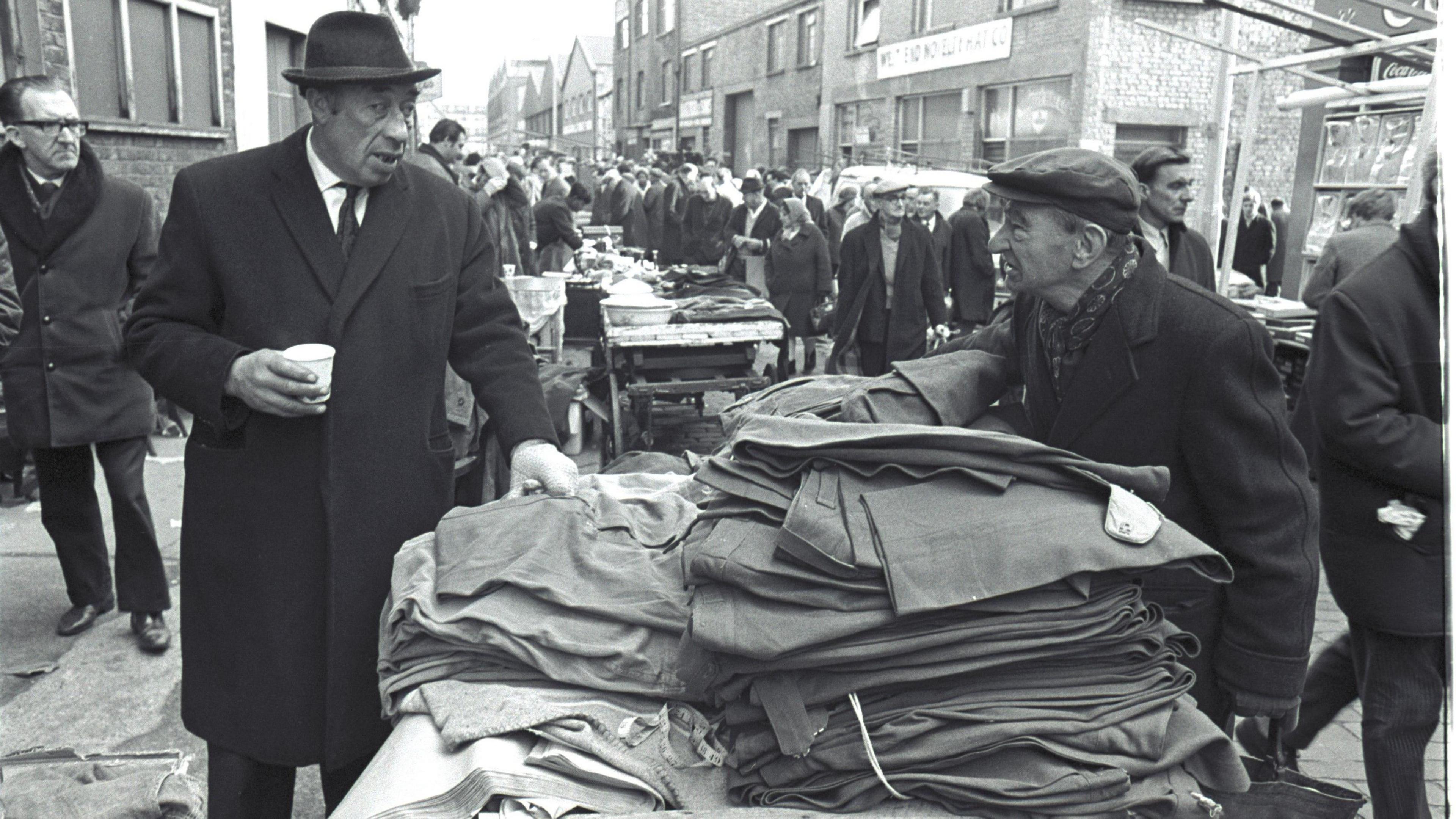 Black and white image of two men in hats and overcoats at an outdoor market. They are chatting over a pile of trousers on a market stall.