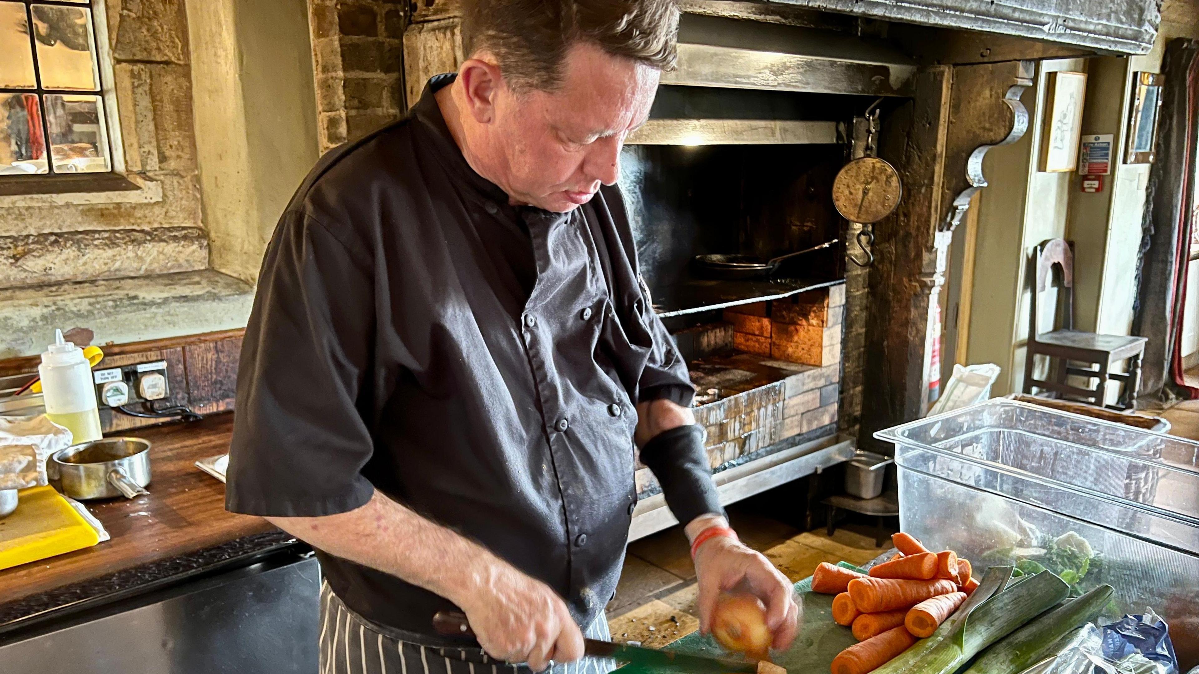 Tony Henderson is in chef's clothing. A black chef's jacket and a striped pinny. He is standing in front of an open fireplace where they cook meat at the Gunton Arms near Cromer. He is standing in front of a green chopping board and is chopping onions. Tony often works in a customer facing position cooking meat. His left arm has an elasticated armband on it to protect his arm where the transfusion fistula goes into his arm.