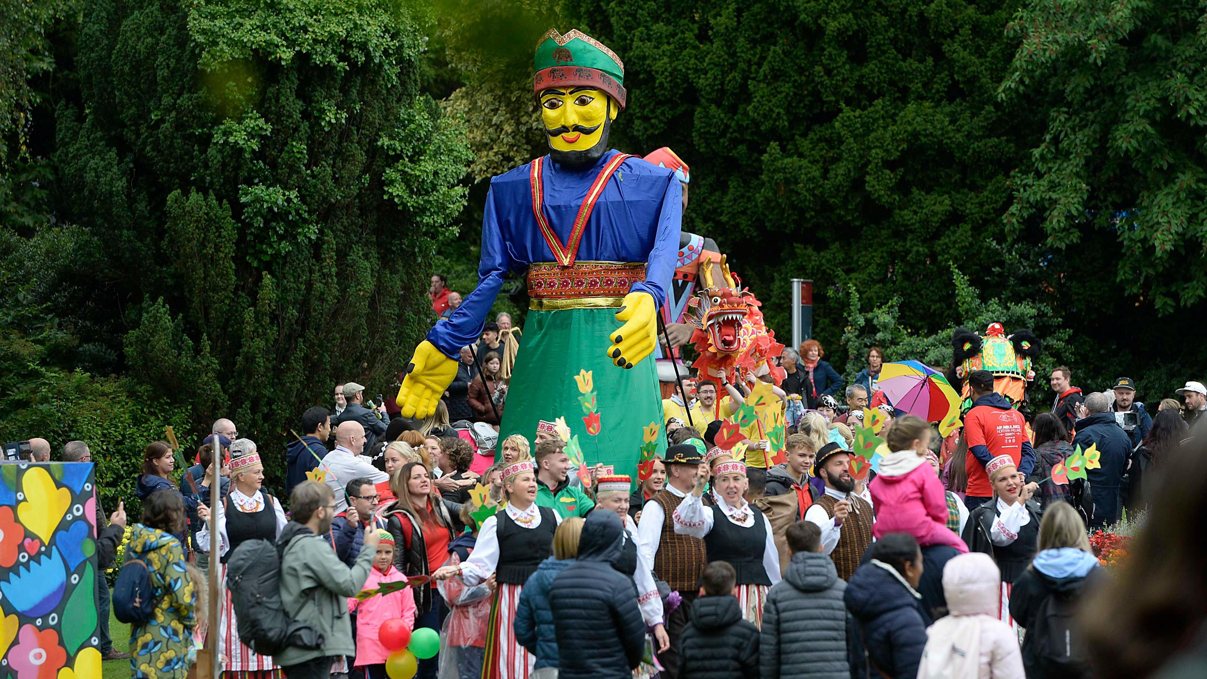 Large puppet being carried through Botanic Gardens by group of people, some in their national dress, as part of Mela day