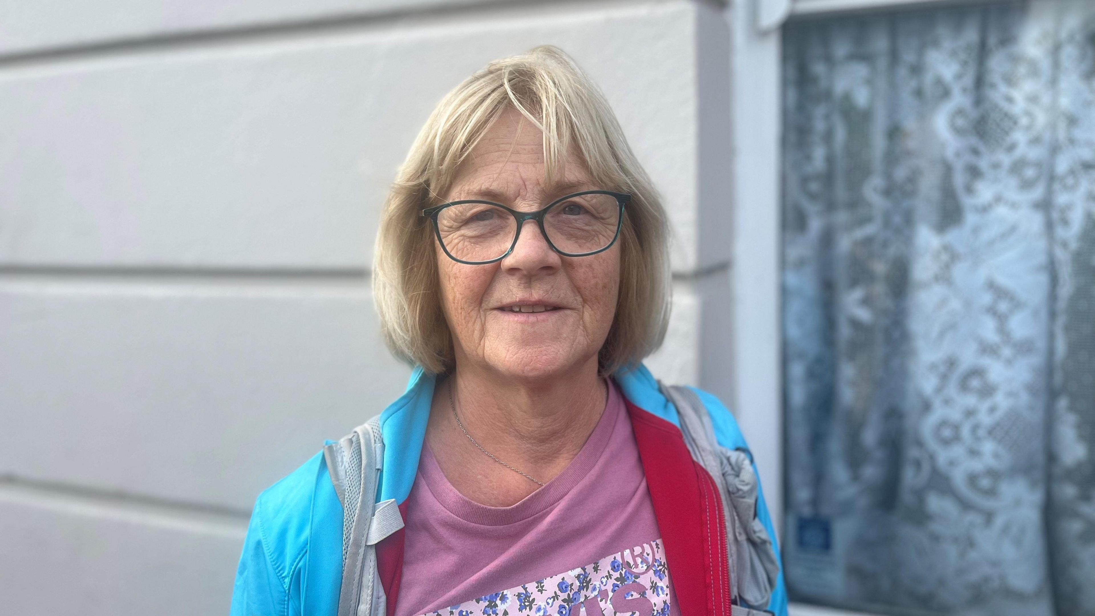 a blonde woman wearing a blue coat with pink top, she is wearing glasses.