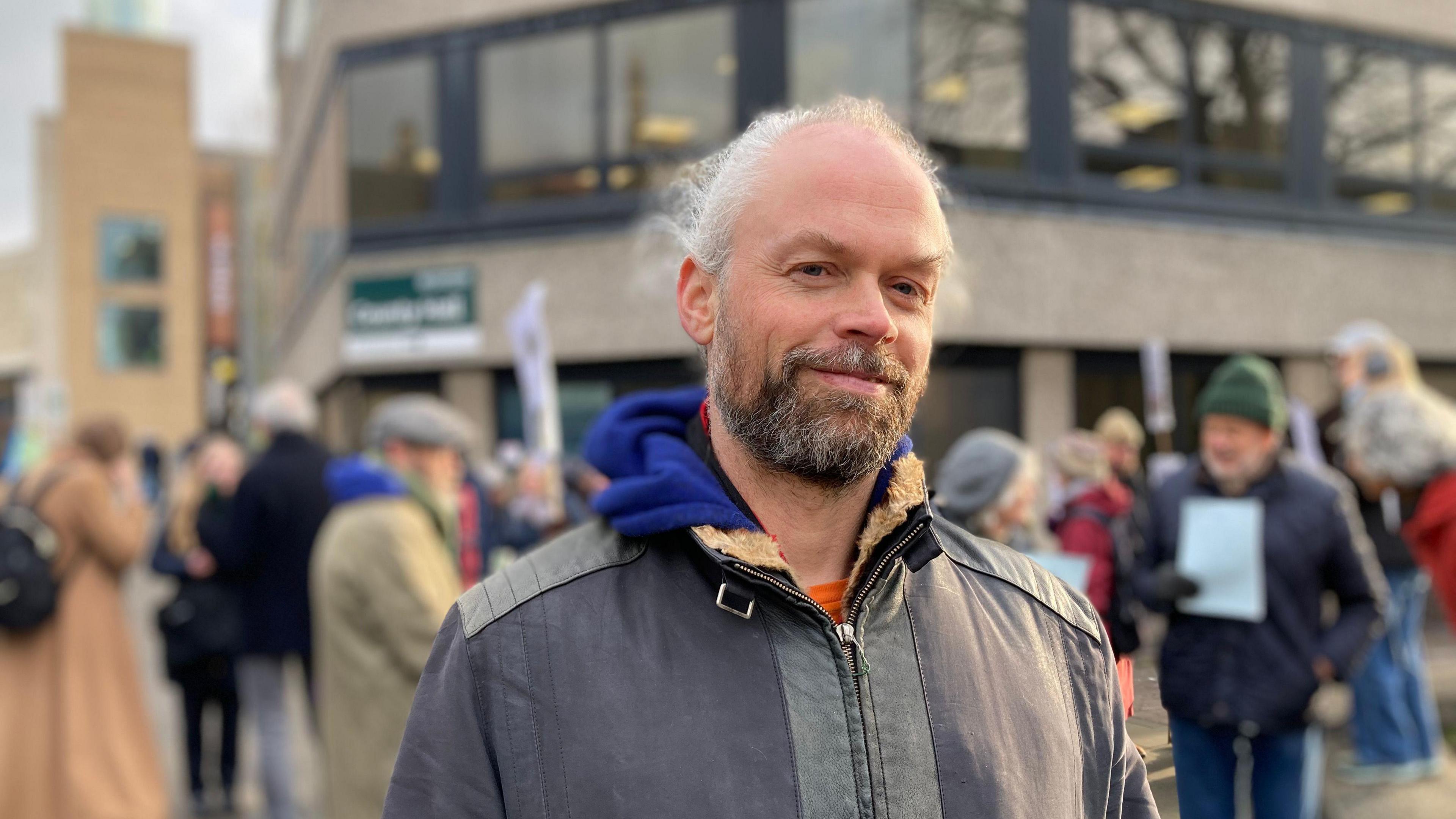 Campaigner Dan Glazebrook poses for a photo outside County Hall in Oxford with protesters in the background. He is balding with wispy white hair, and a brown beard. He is wearing a grey jacket with a blue hoodie underneath. 