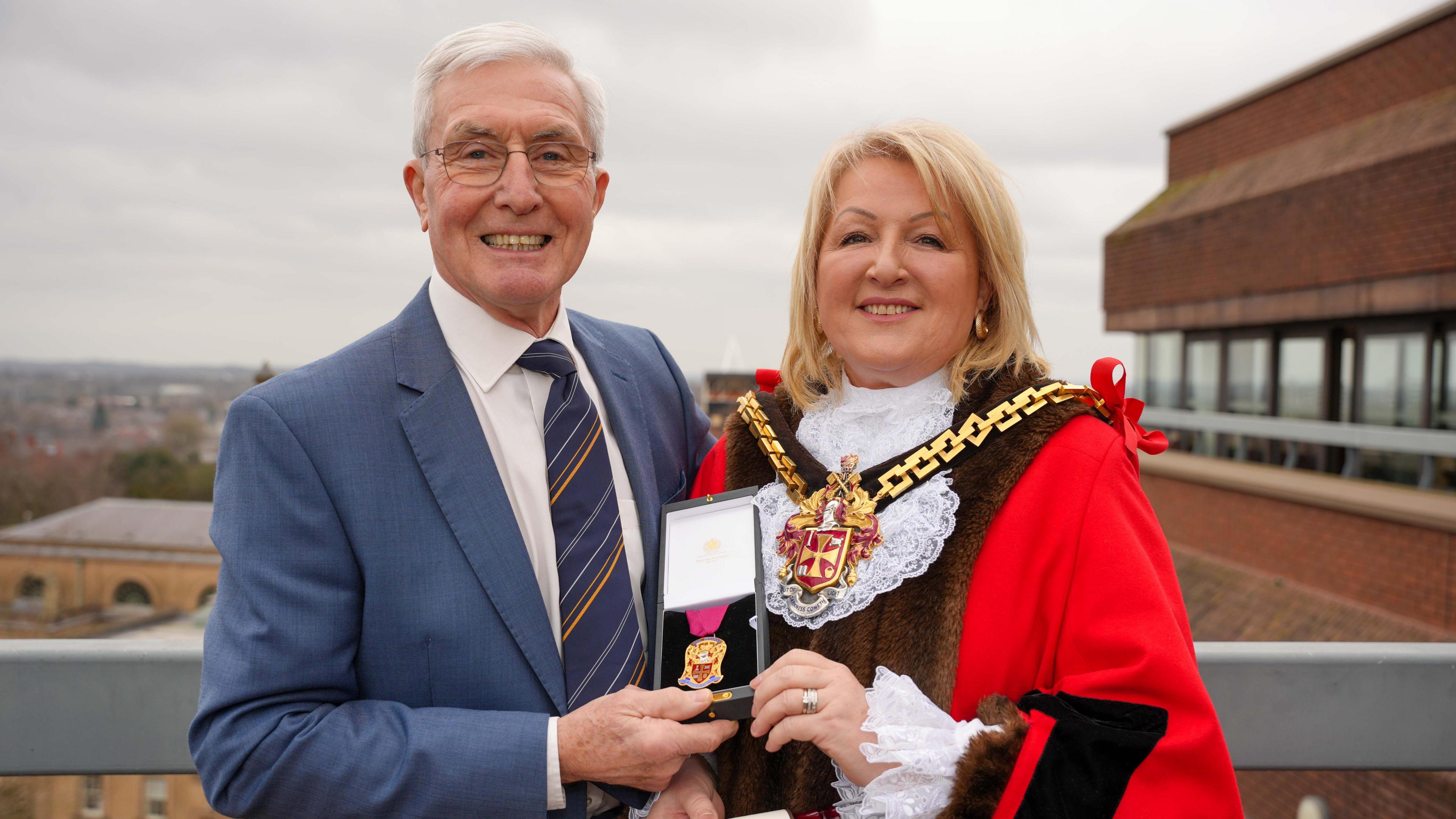 An older man in a blue suit and white shirt standing next to a lady in a red cloak with brown fur. The lady is wearing a gold medallion and between them they are holding a smaller gold medal. 