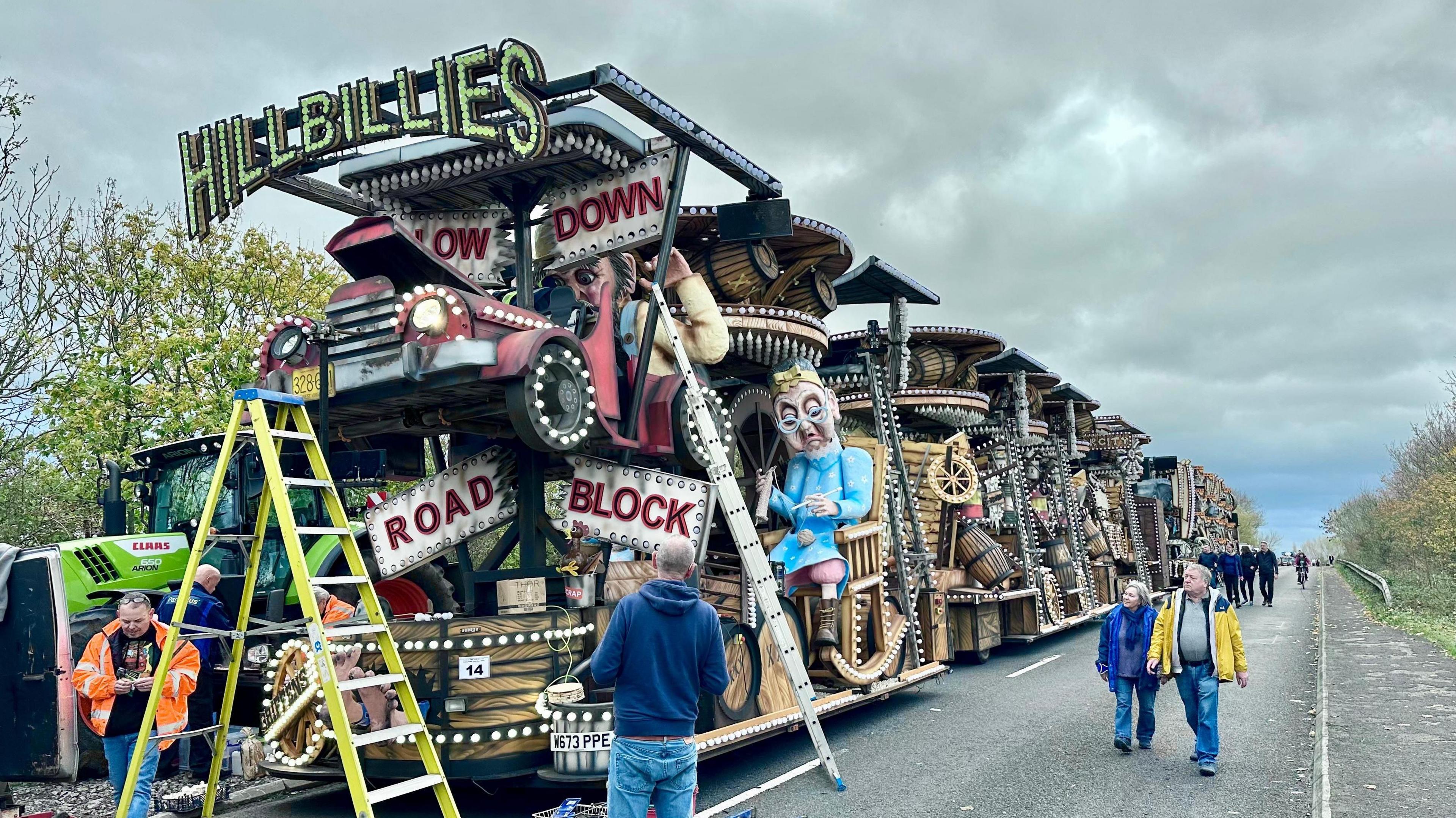 A huge Somerset carnival cart sits at the front of a line of other carts on a main road. There's a ladder up on one side with repairs taking place. There's a few people looking at the carts.